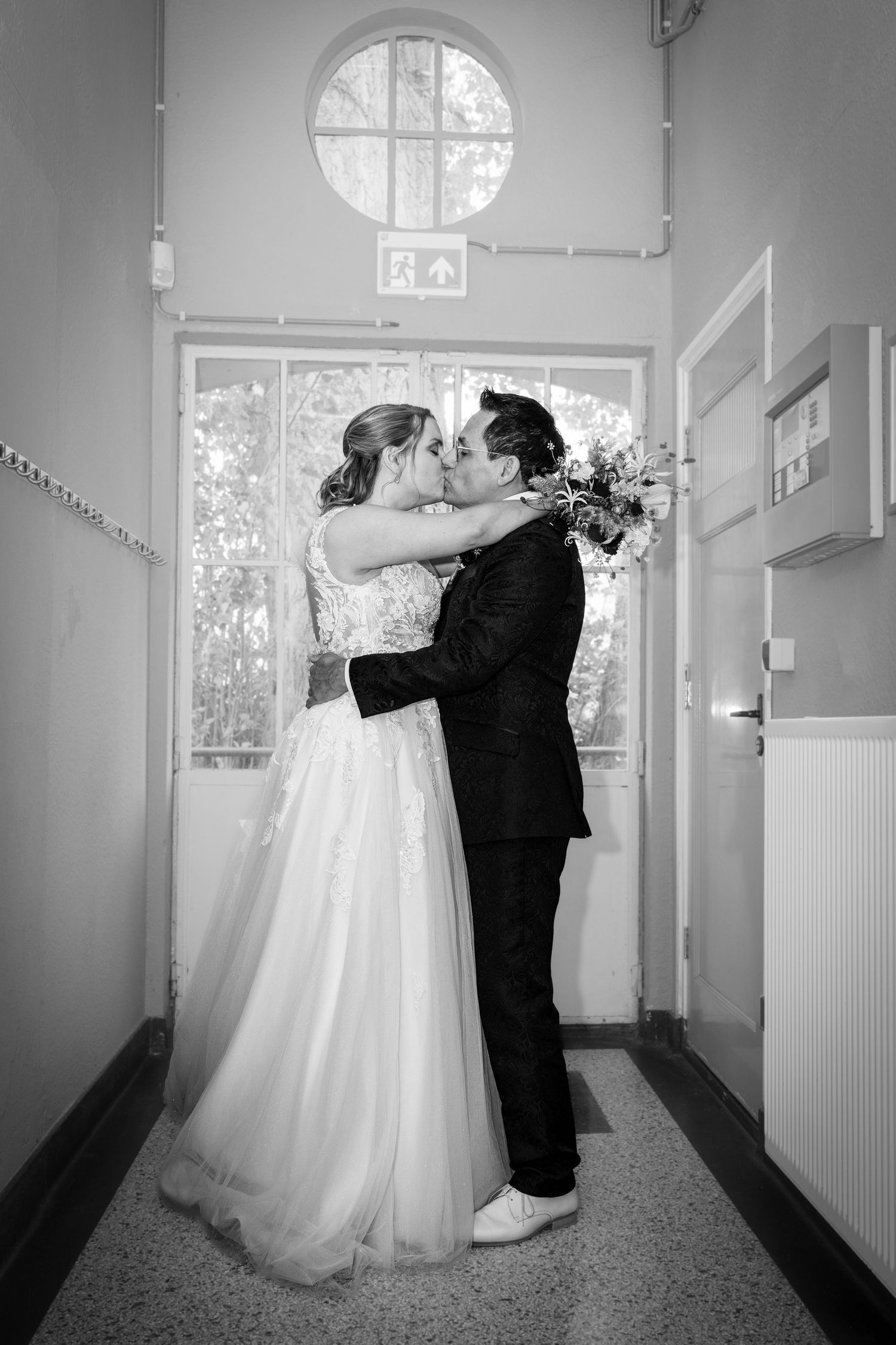 Black and white photo of newlyweds embracing in doorway with round window above.