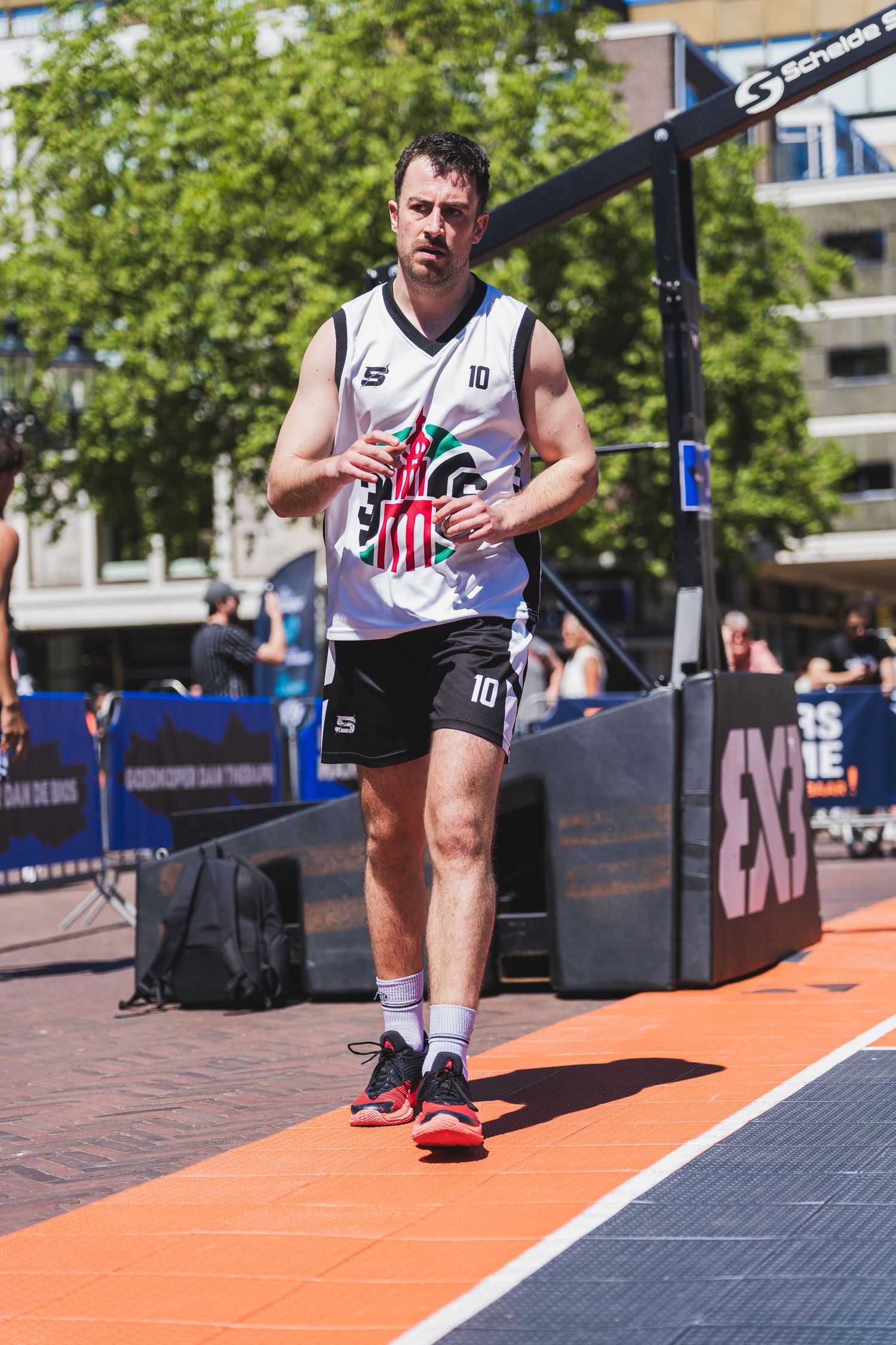 A male basketball player wearing jersey number 10 walks on an outdoor 3x3 street basketball court on a sunny day.