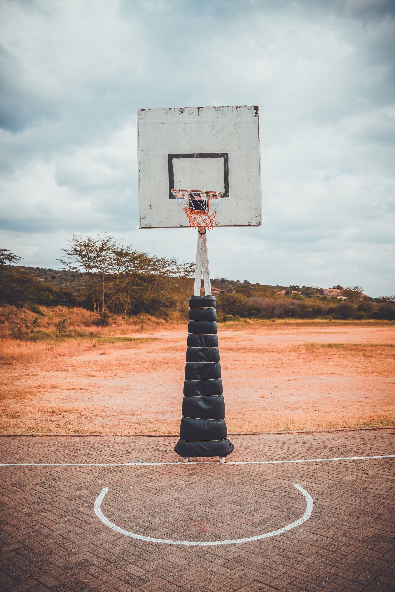 Outdoor basketball hoop with padded pole on a brick court against a cloudy sky in a rural African setting.