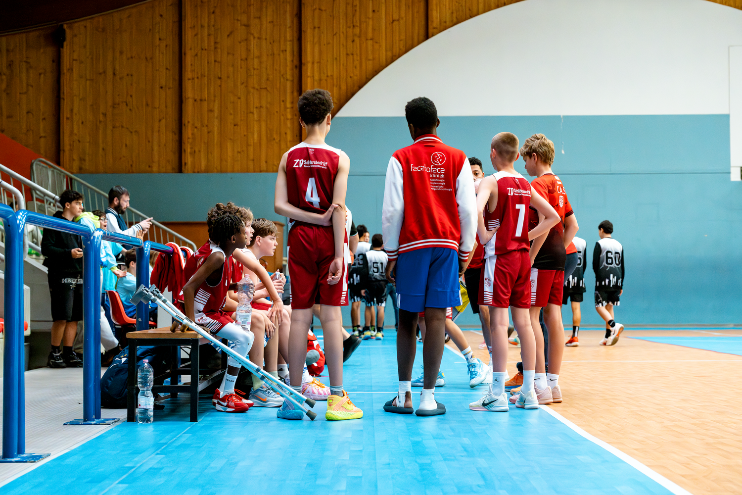 Youth basketball players in red jerseys standing on sideline during indoor game.