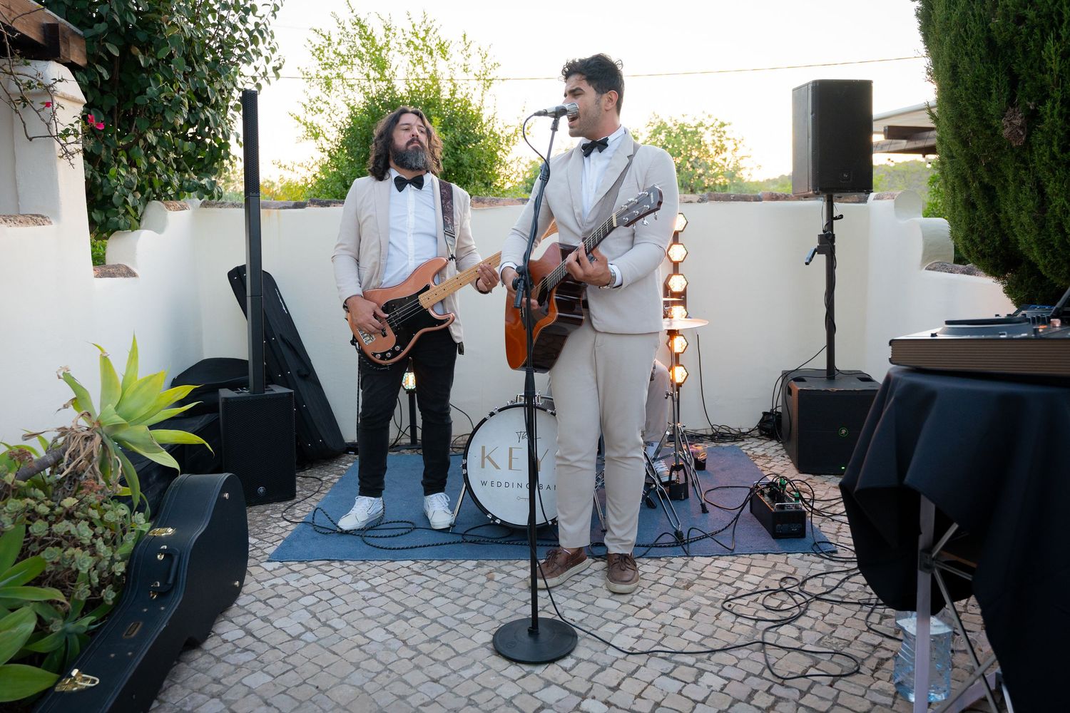 Musicians perform with guitars and microphones at an outdoor patio venue with speakers and sound equipment set up.