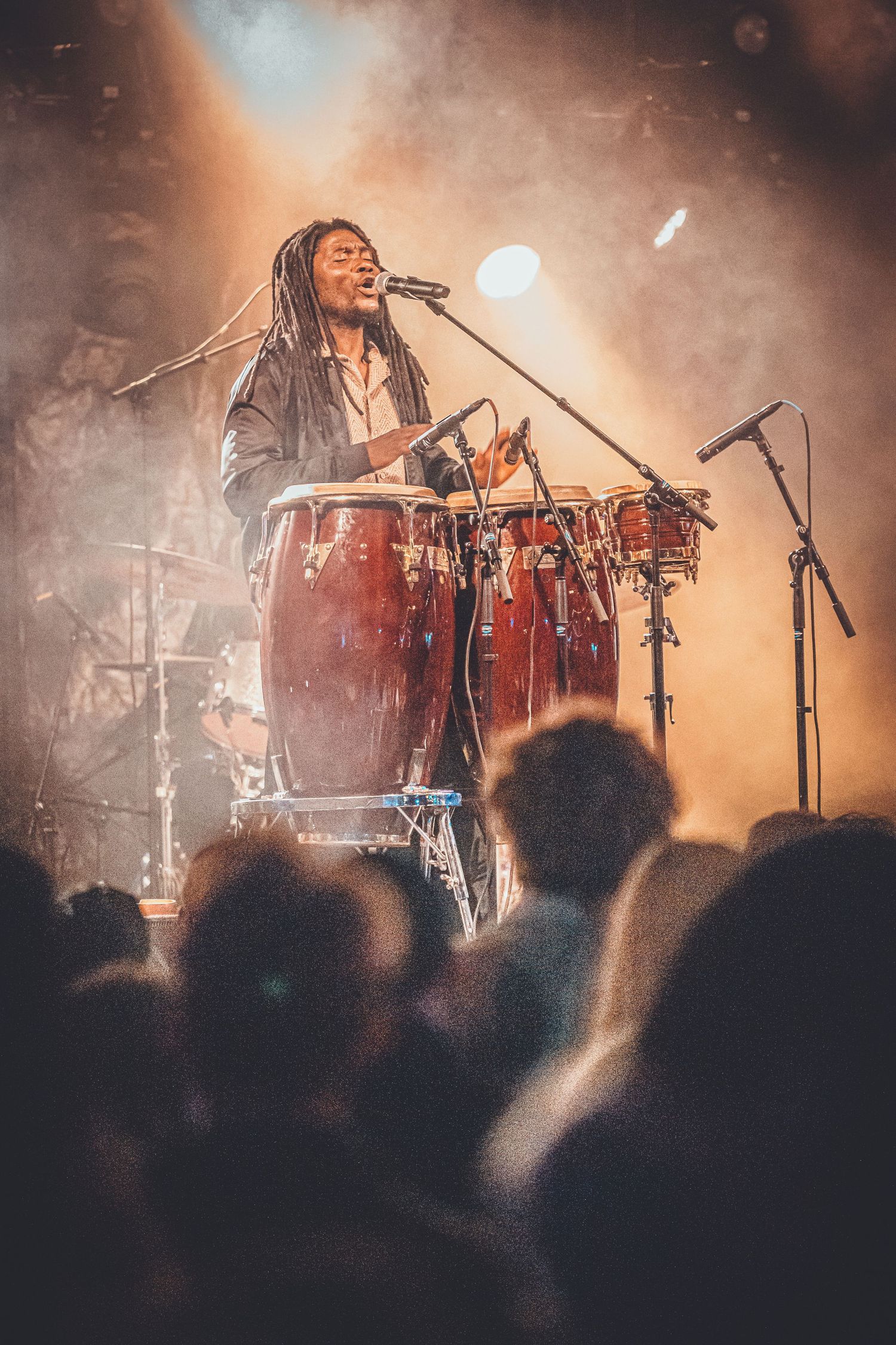 Musician playing congas on stage during atmospheric performance with moody lighting and crowd silhouettes in foreground.