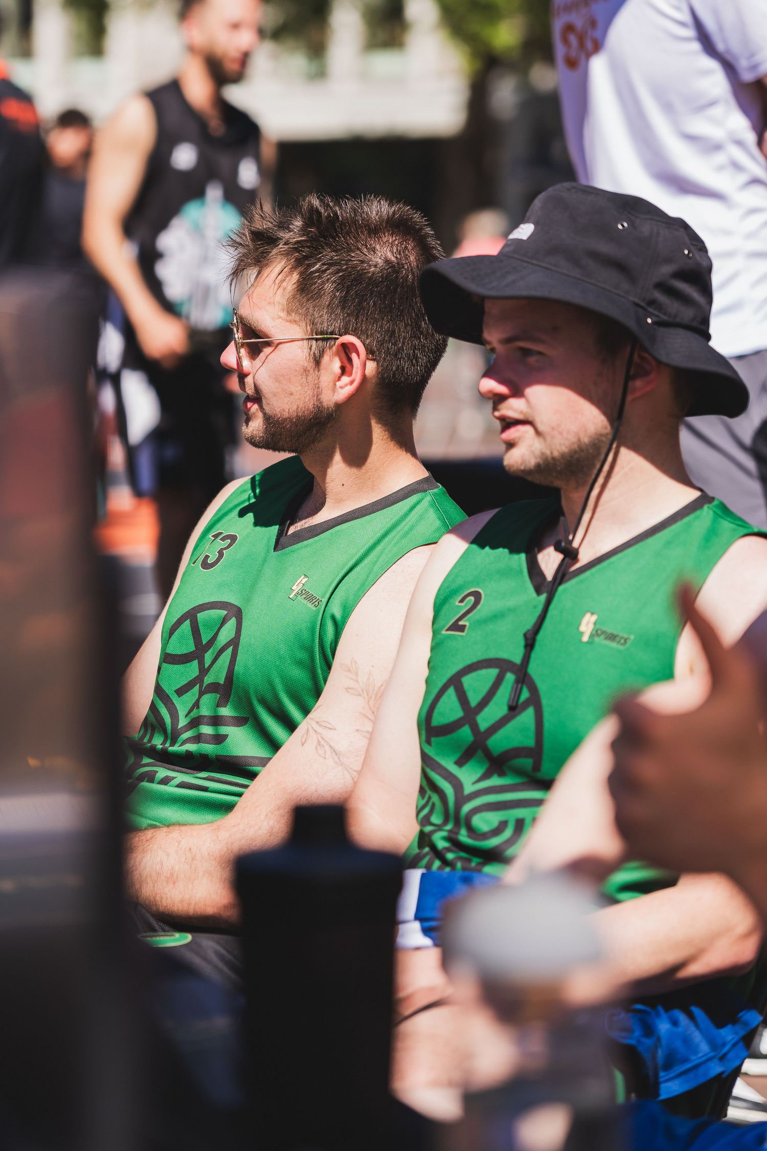 Two men in green numbered basketball jerseys watch an outdoor 3x3 basketball tournament on a sunny day.