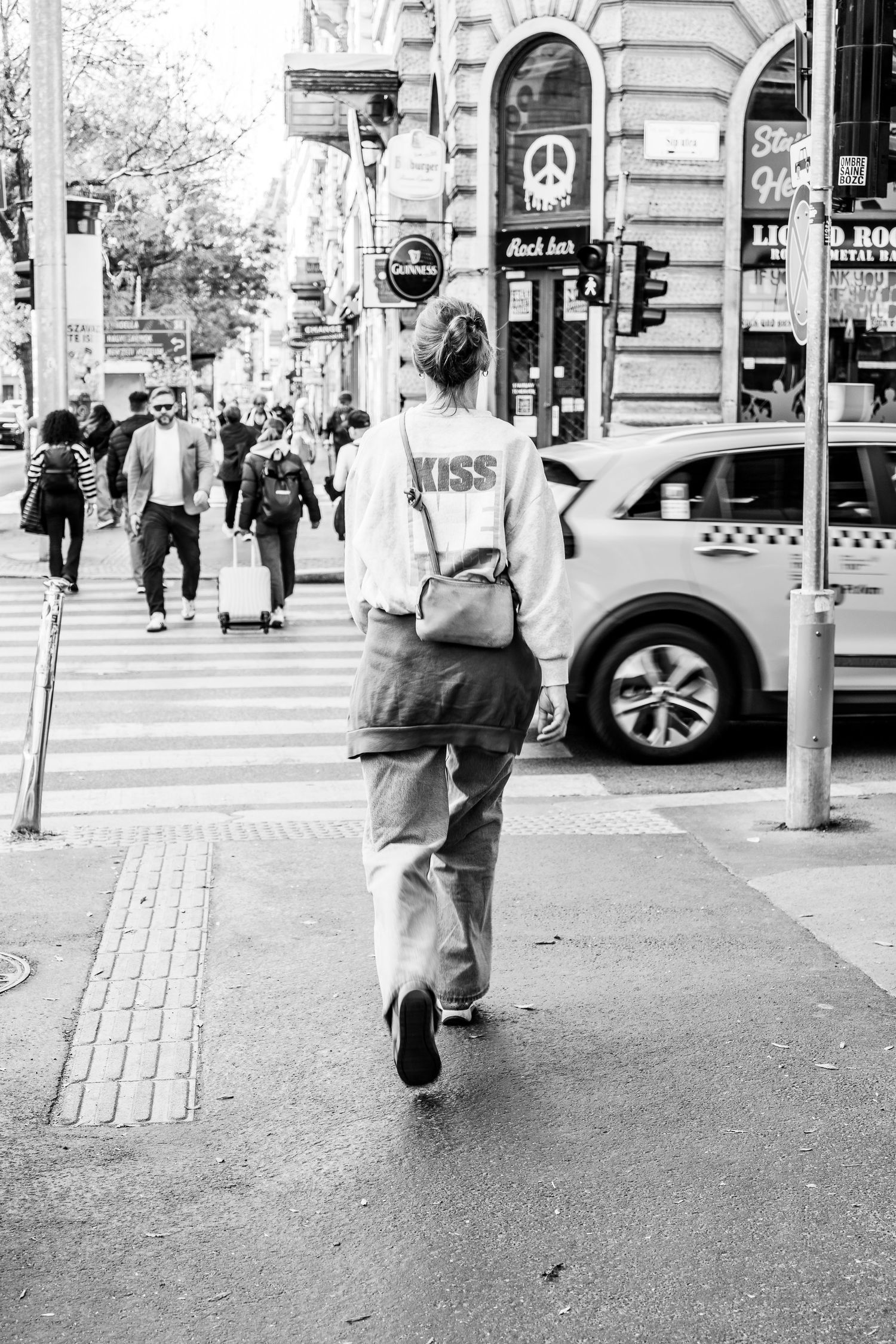 Black and white street photography of pedestrians walking on a city sidewalk with cars and buildings in the background.