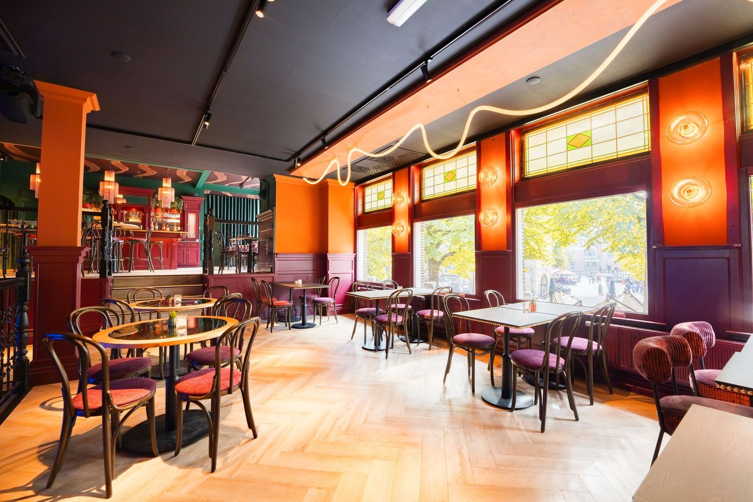 Stylish restaurant interior with orange walls, herringbone wood floors, velvet chairs, and large windows.