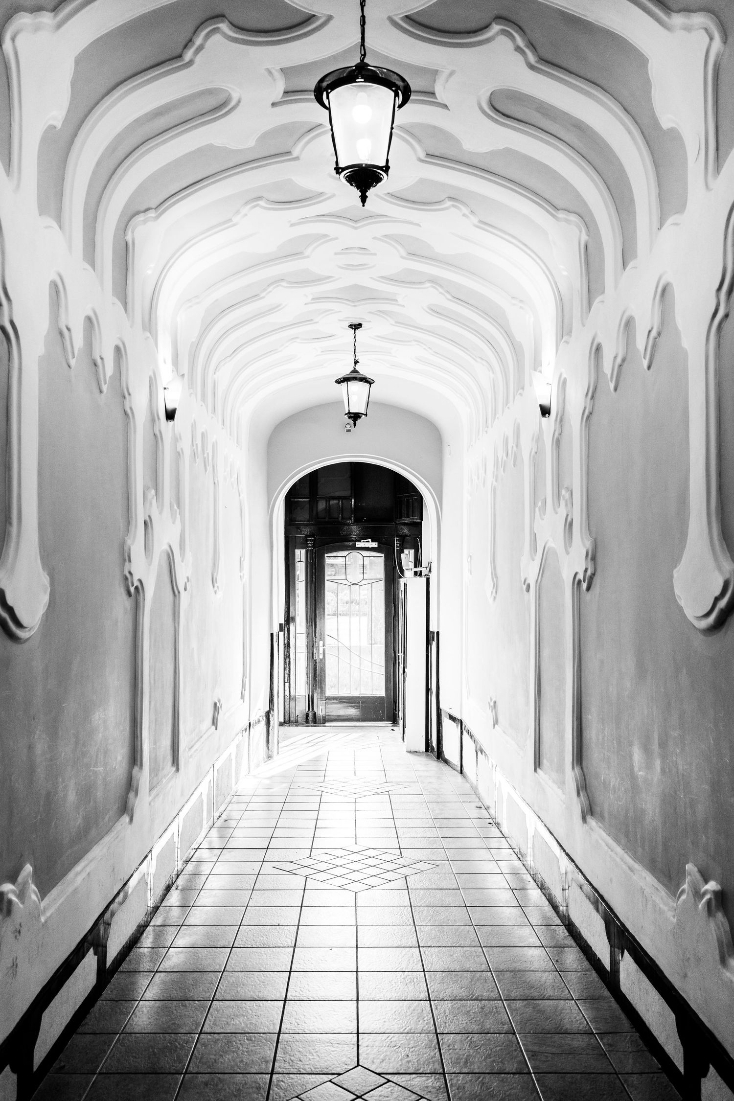 A dramatic black and white perspective of an ornate arched hallway with hanging lamps creating a vanishing point effect.