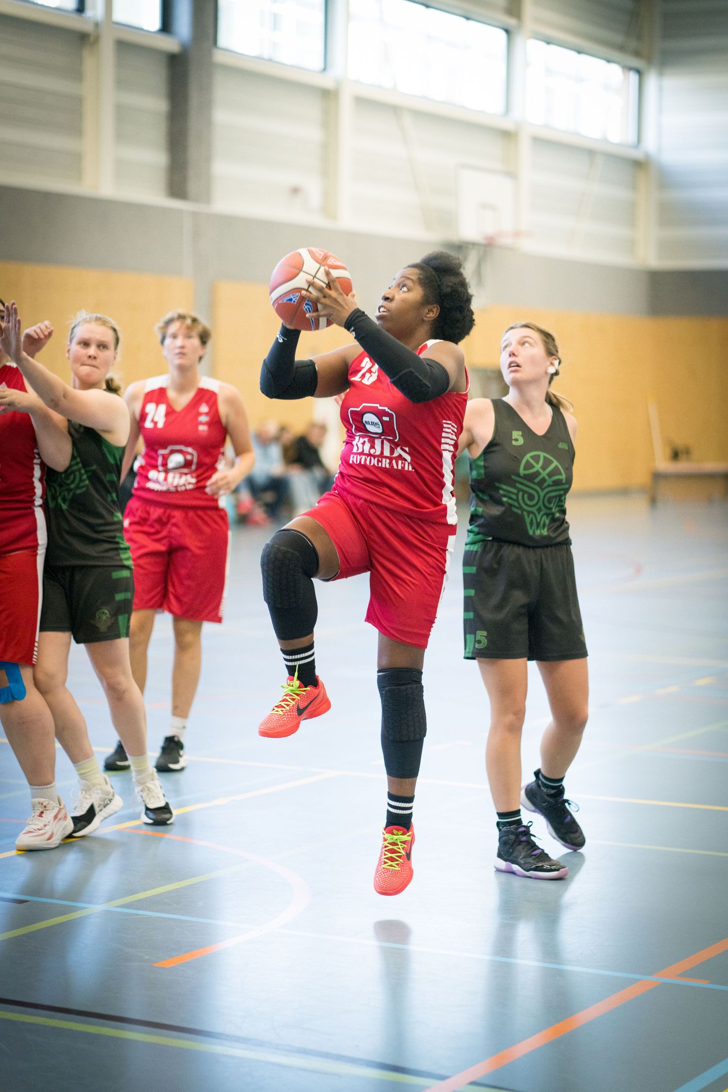 Basketball players compete in an indoor game with one player in red jersey jumping high to make a shot between defenders.