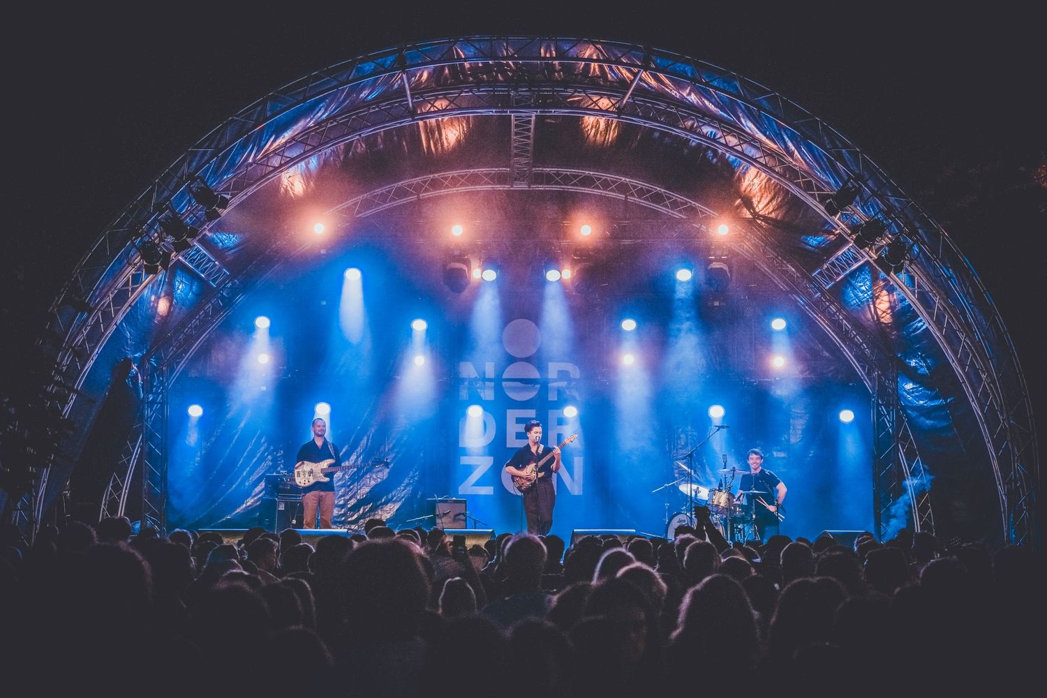 Concert stage lighting creates dramatic silhouettes of performers beneath a curved arch with blue and pink illumination.