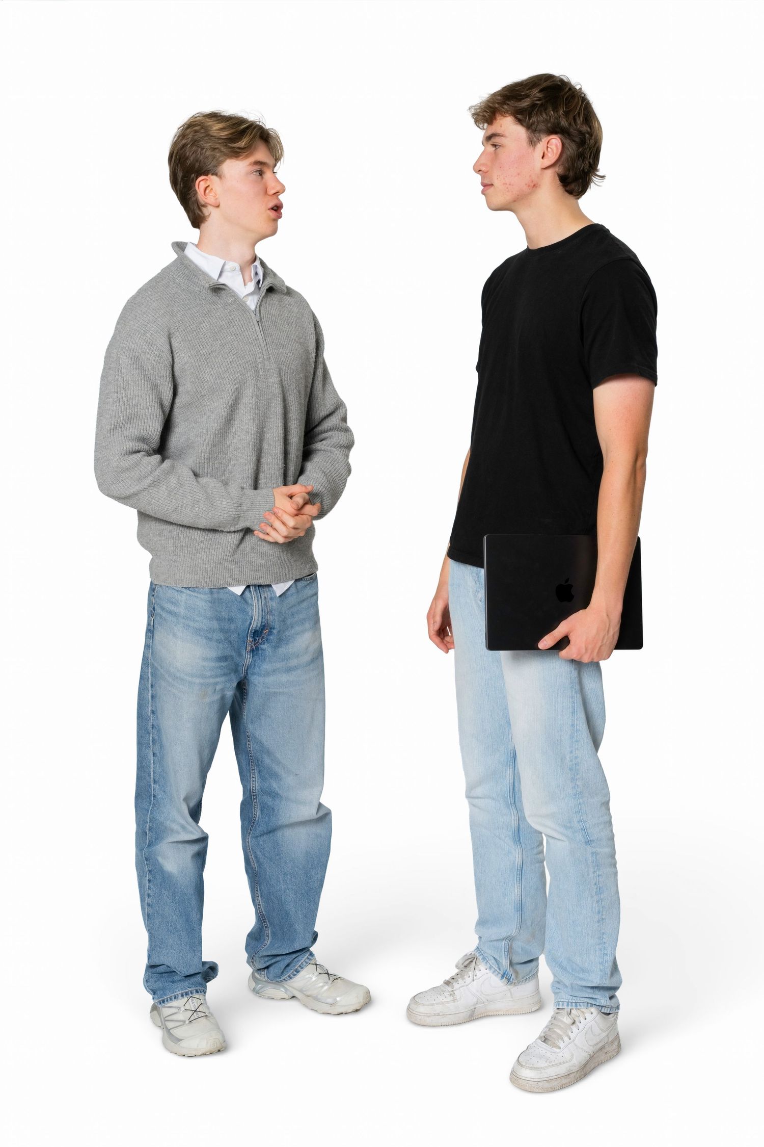 Two teenage boys in casual clothes stand facing each other and talking, one in a gray sweater and jeans, the other in a black t-shirt holdin