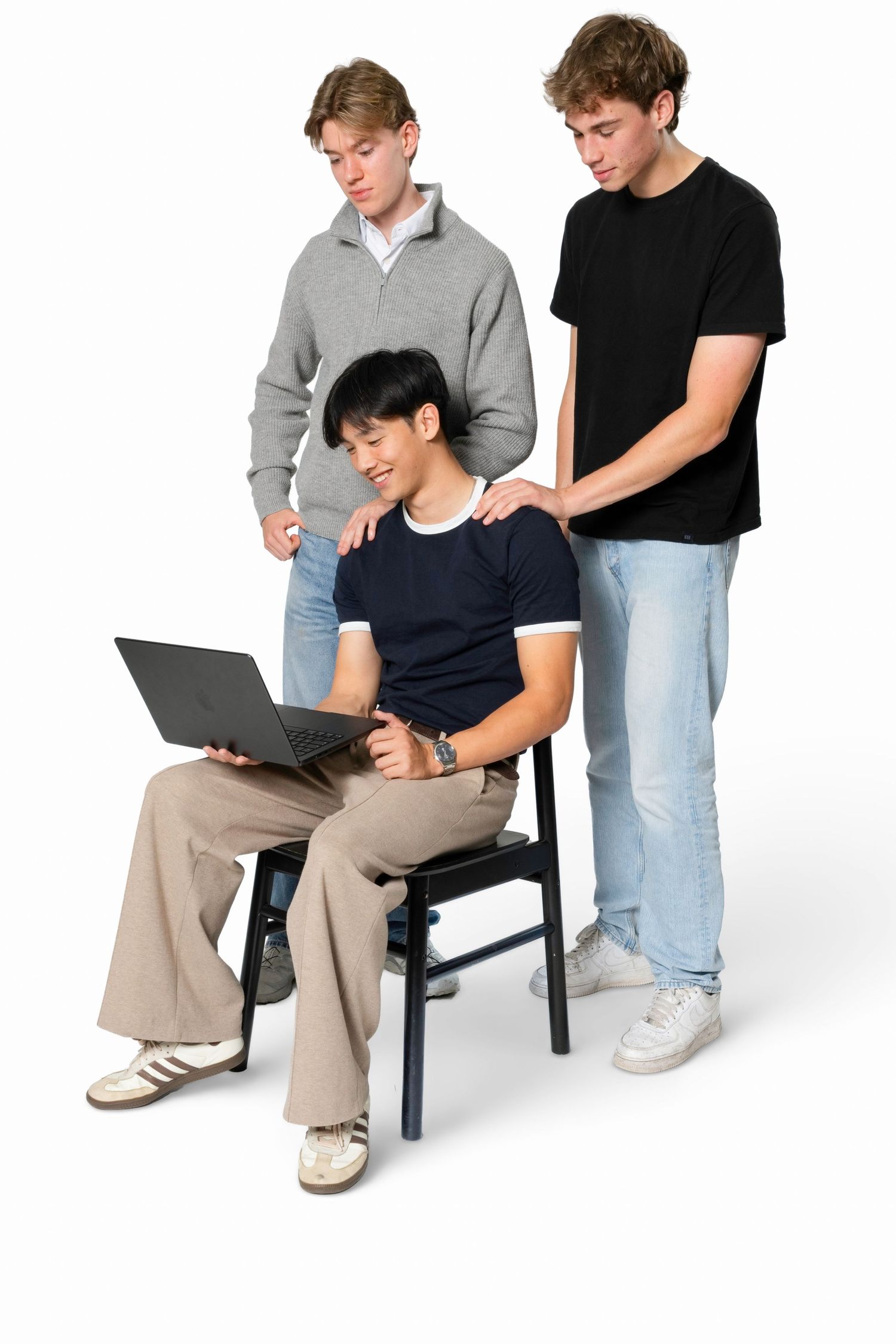 Two young men stand behind a seated friend in a navy t-shirt as they all look at a laptop together against a white background.