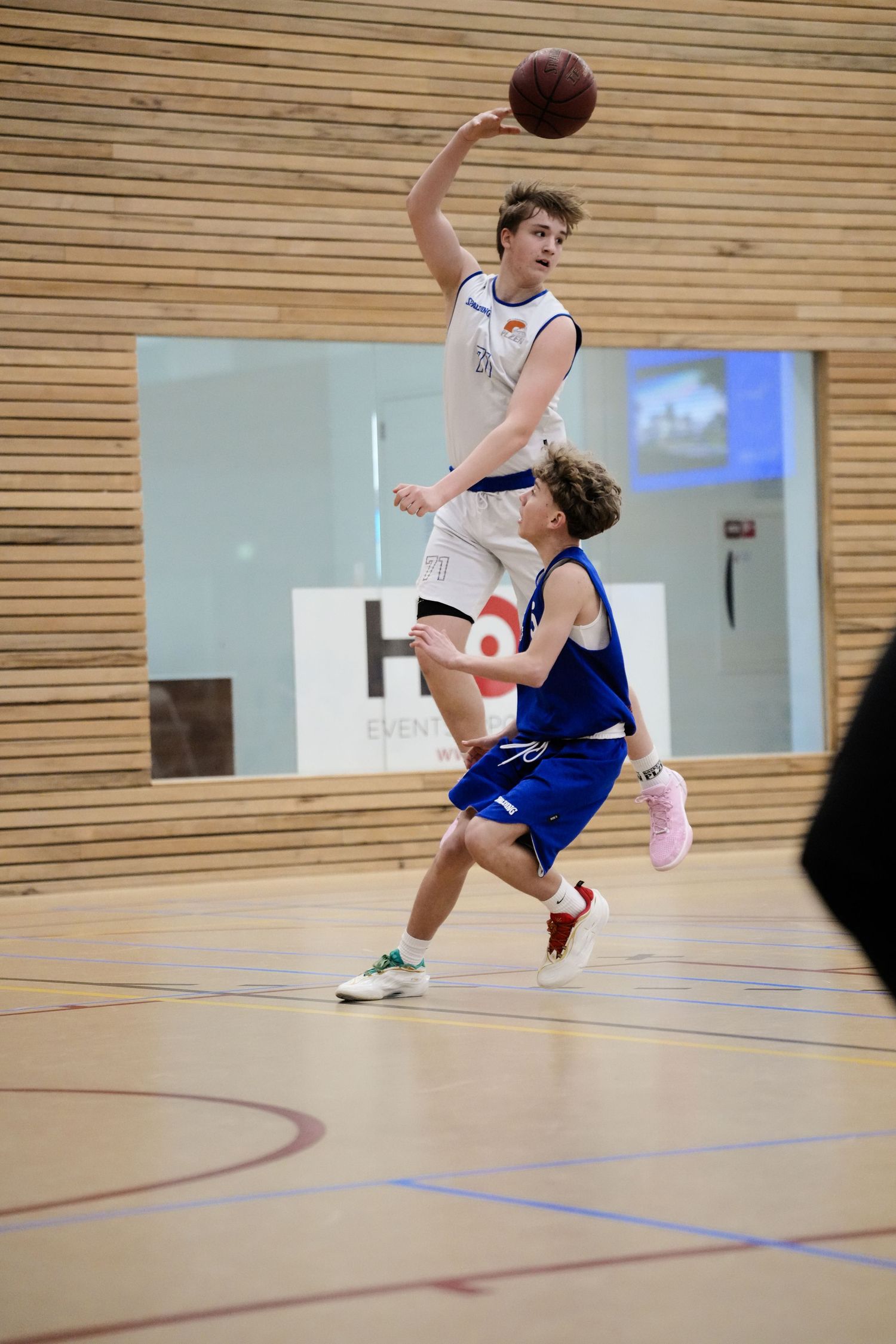 Two youth basketball players compete indoors, one in white leaping to shoot over a defender in blue uniform.