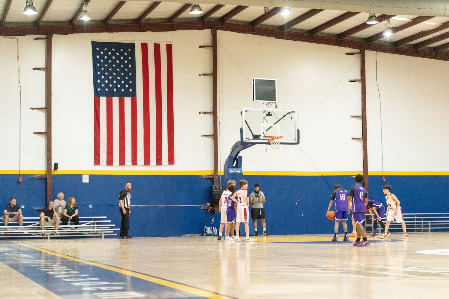 Indoor basketball game in a gymnasium with an American flag on the wall and players on the court.