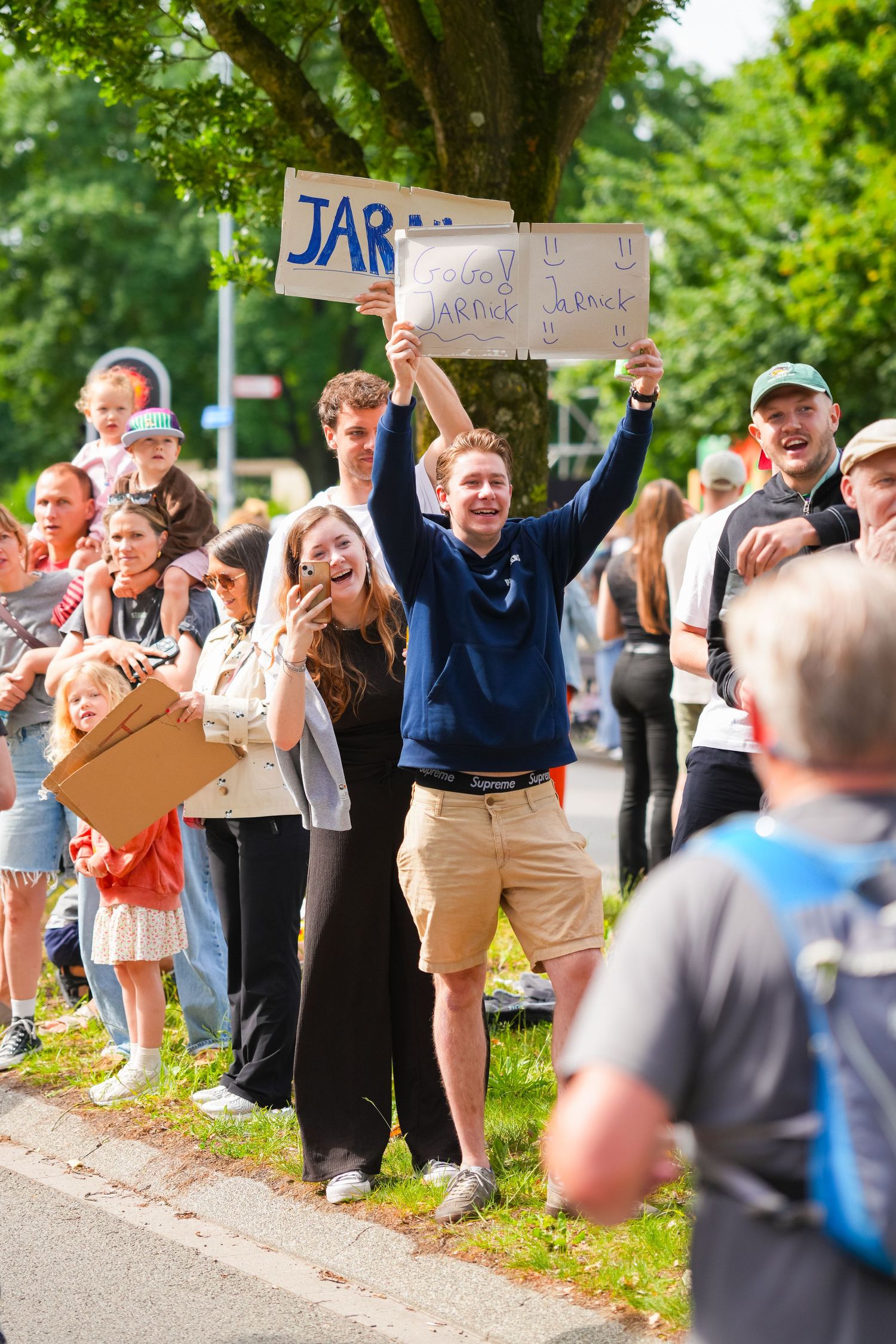 Excited crowd cheering at outdoor event, man holding JAR sign above head, green trees in background.