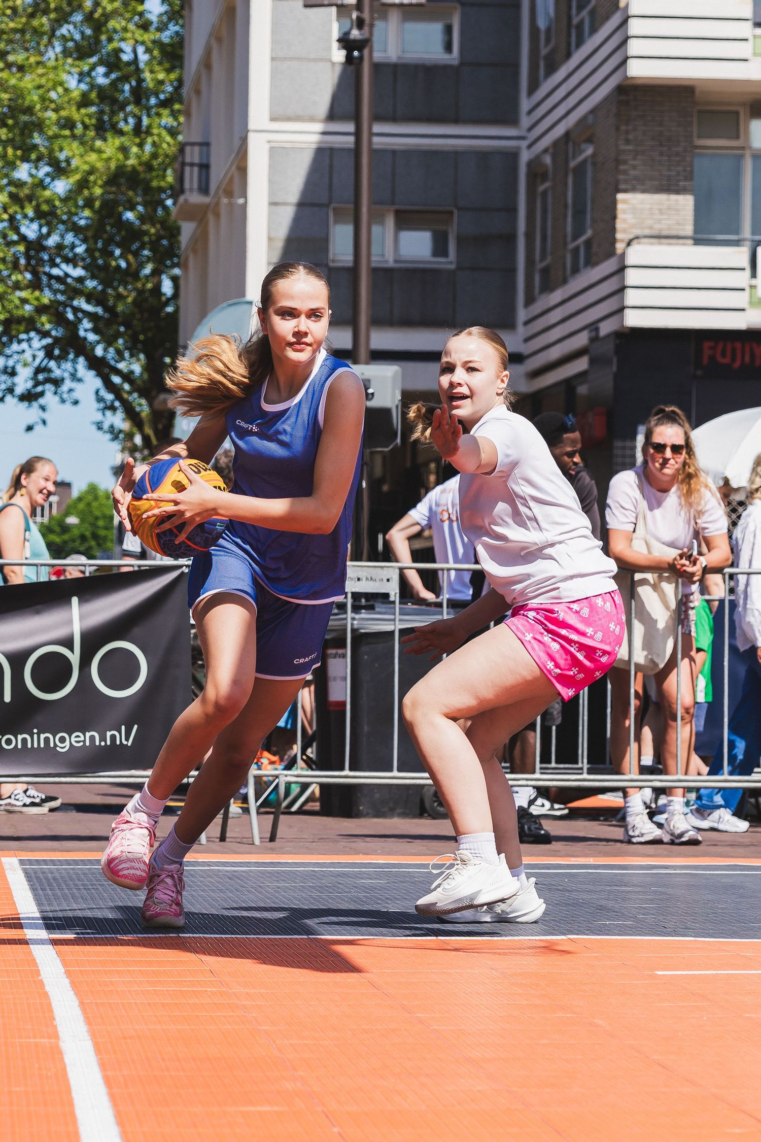 Two girls playing street basketball outdoors, one in blue holding ball, other in pink defending, spectators watching behind barriers.