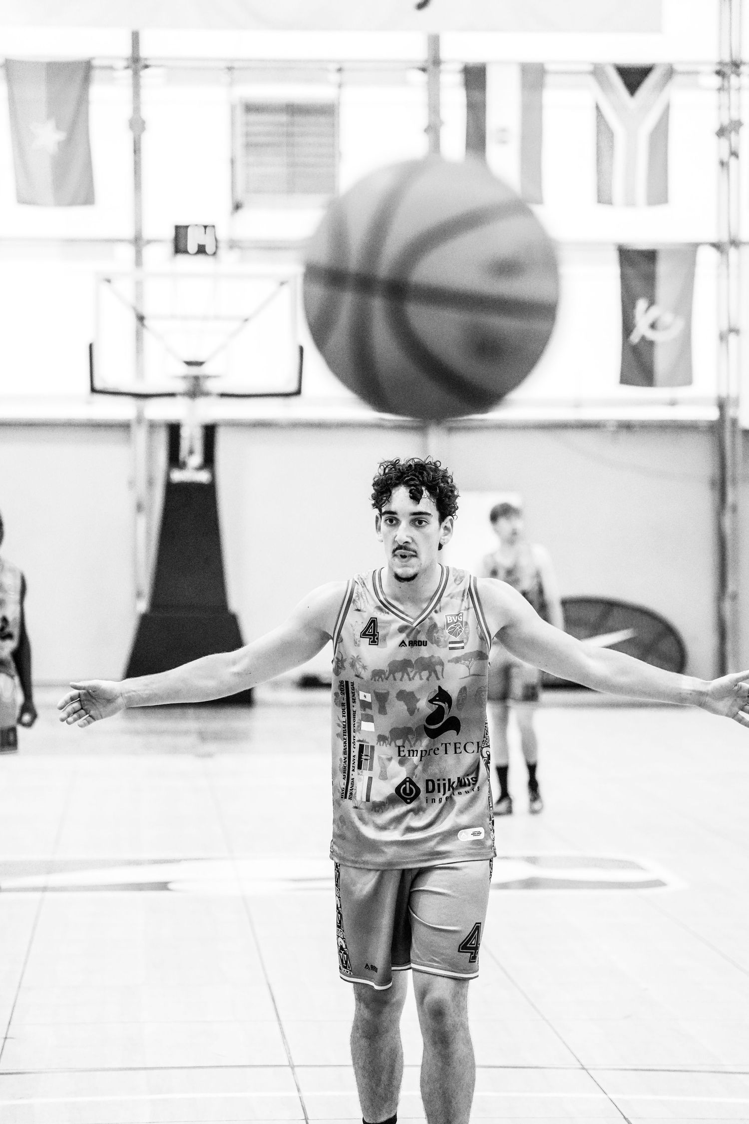 Black and white photo of a basketball player in jersey number 4 with arms spread wide as a ball floats above in a gym.