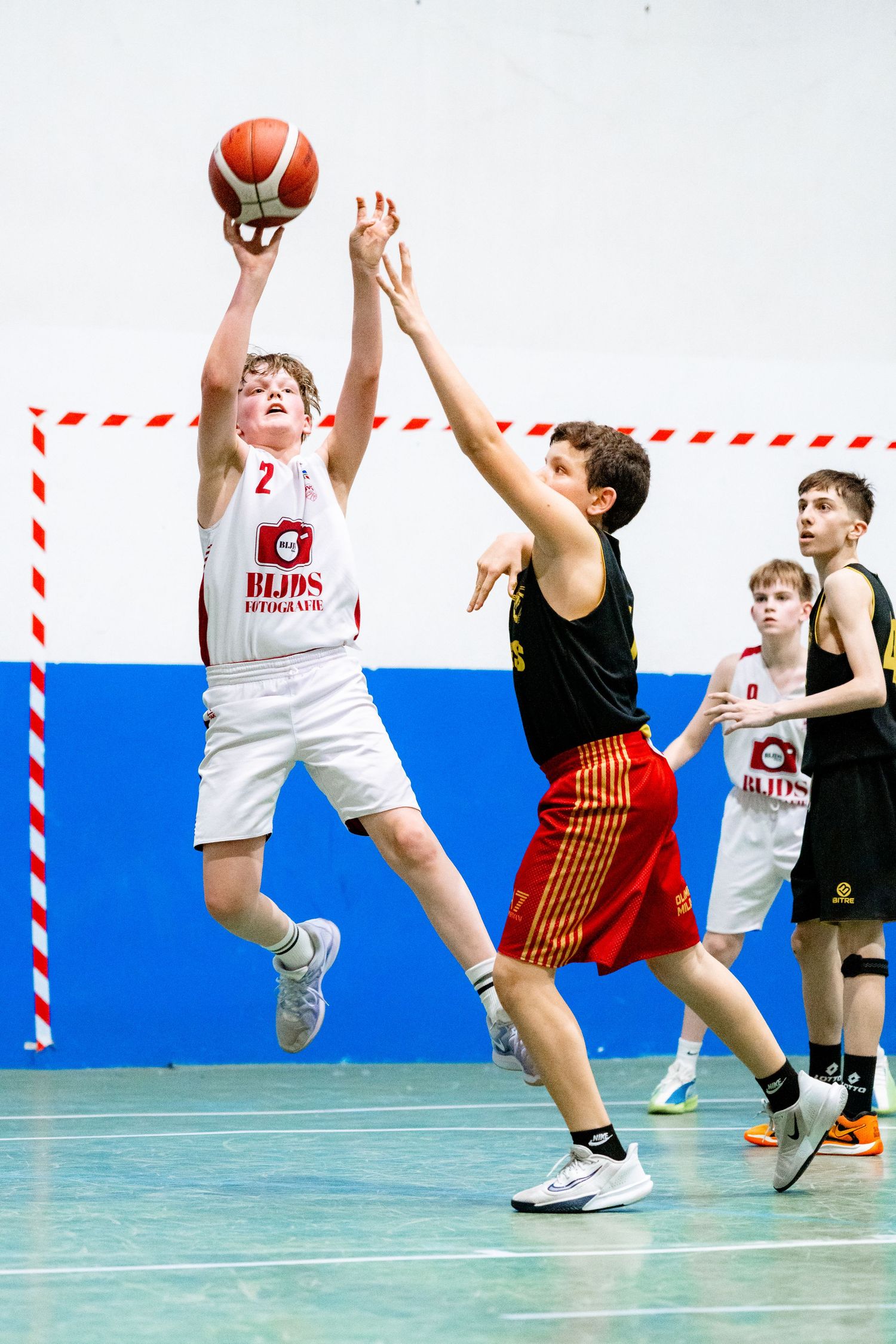 Young basketball player in white jersey jumps to shoot over defender during indoor youth basketball game.