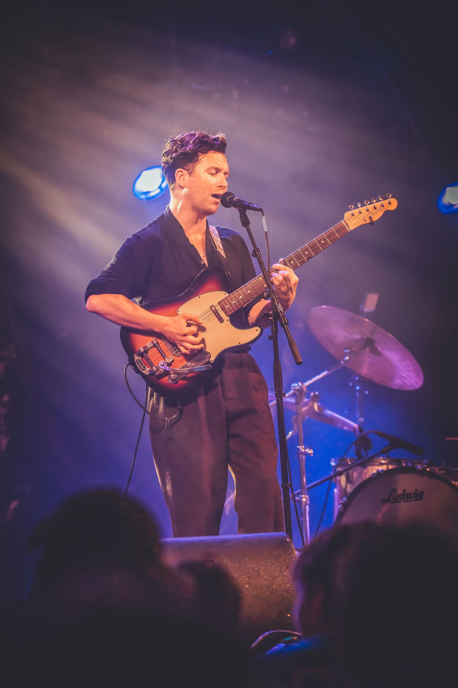 A performer plays an electric guitar on stage under dramatic purple lighting with a drum kit visible in the background.