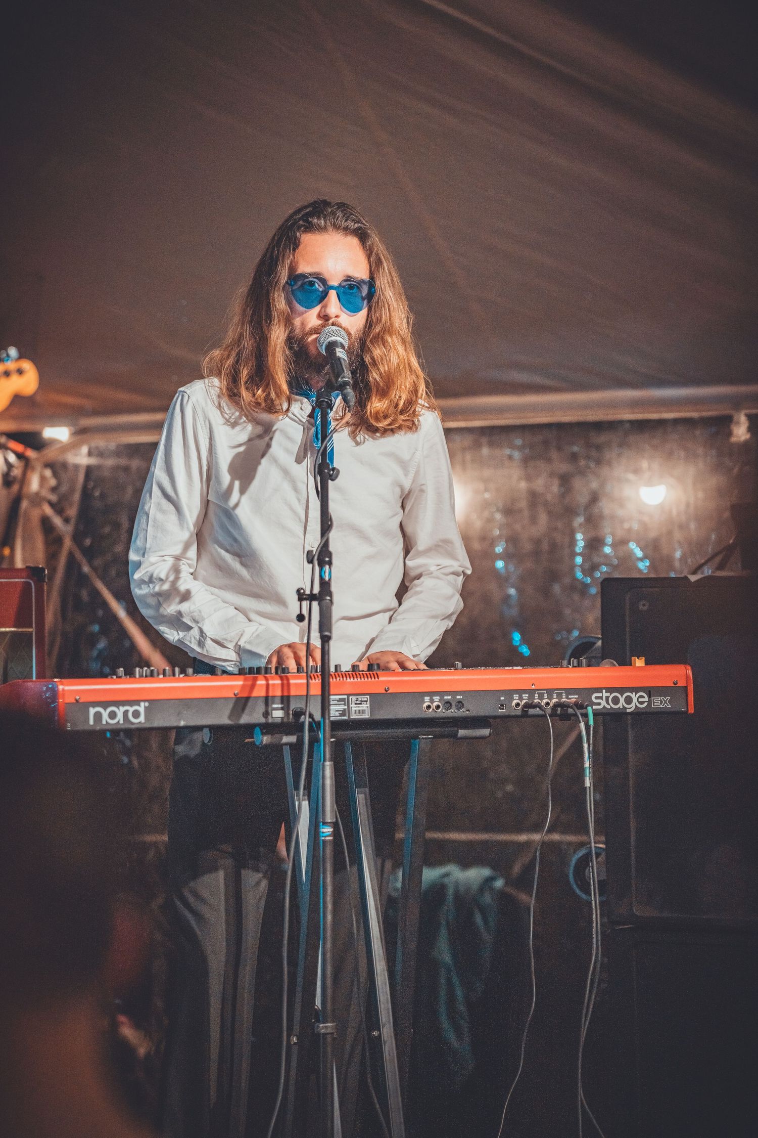 A musician with long hair and blue sunglasses performs at a keyboard during a live concert.