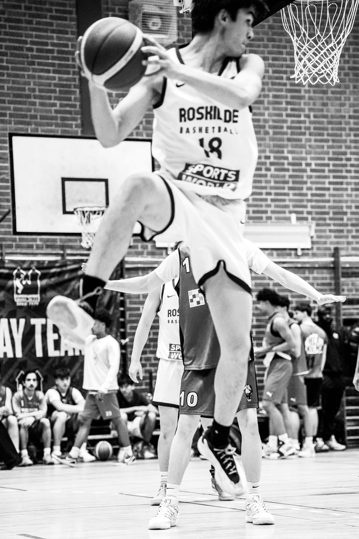 Black and white photo of basketball players in action during an indoor game with spectators watching from sidelines.