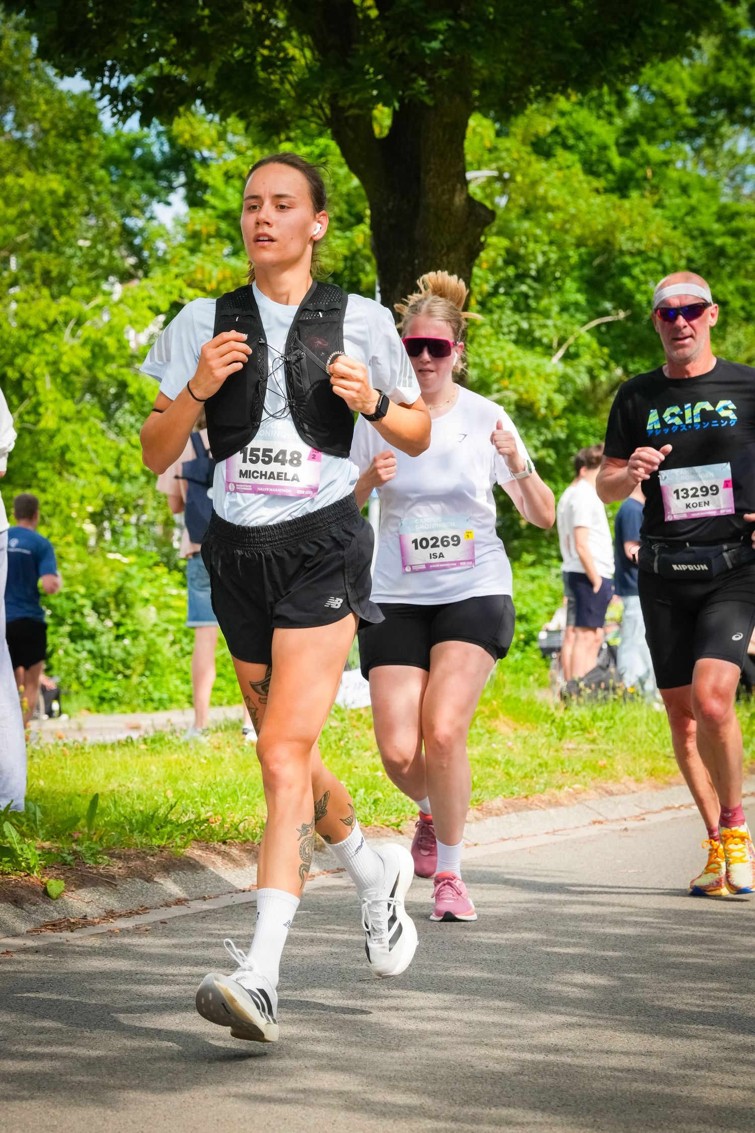 Runners competing in a road race, wearing numbered bibs, jogging through a tree-lined path on a sunny day.