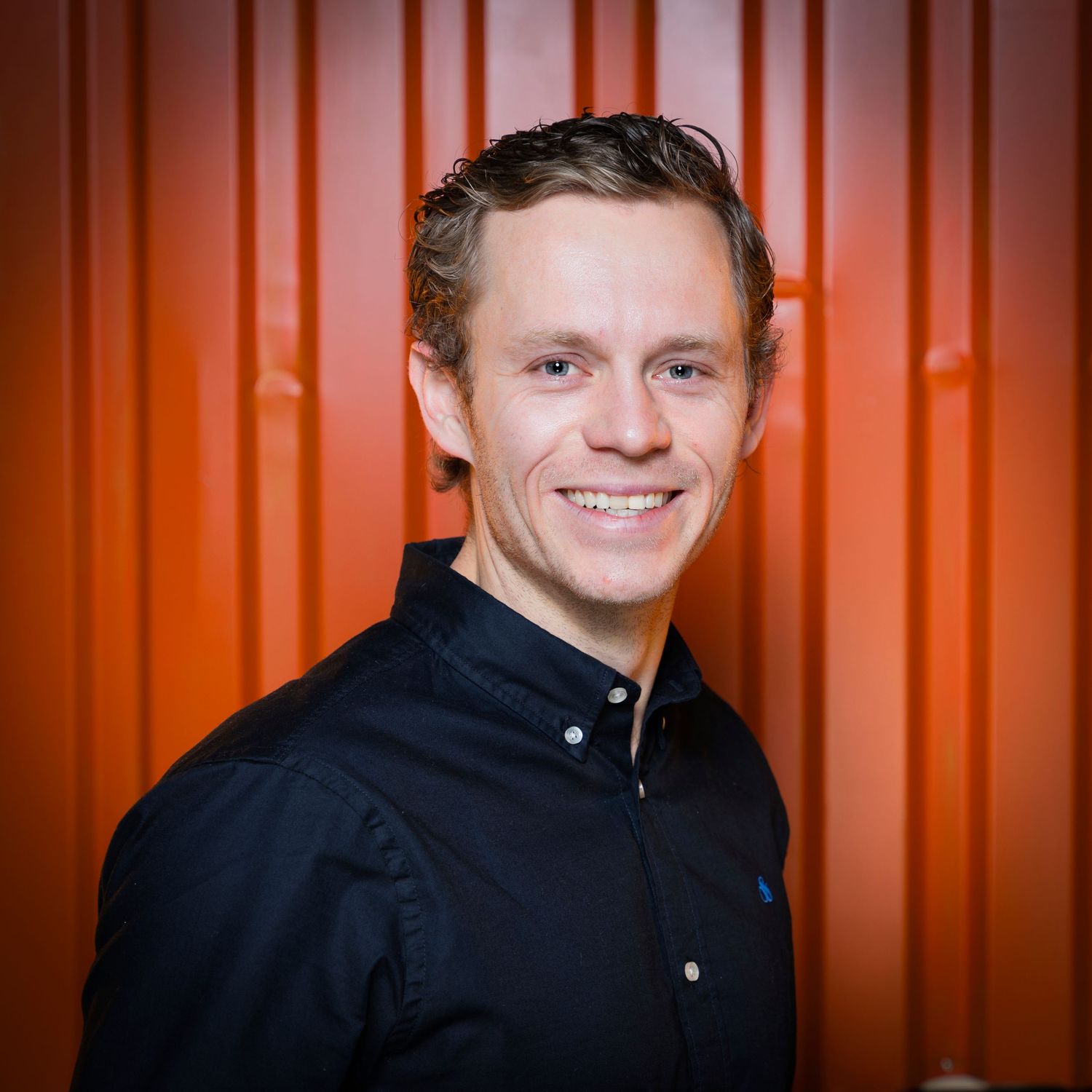 Smiling young man in black shirt poses against vibrant orange wall in professional headshot photo.