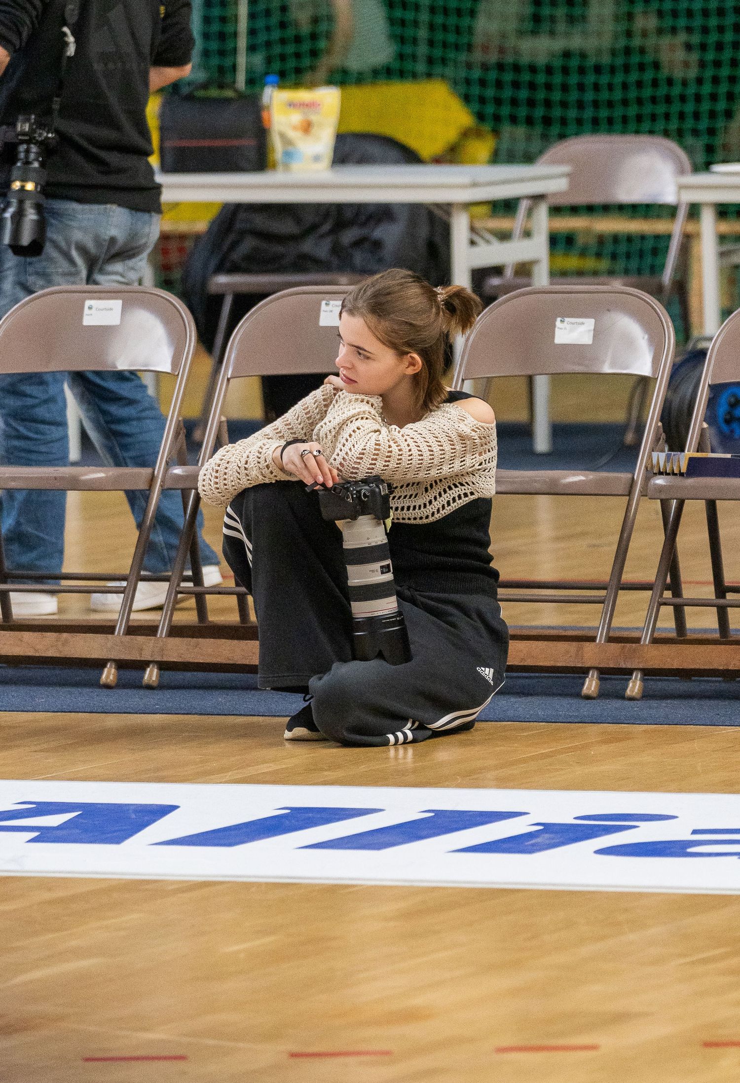 A person in a striped sweater sits cross-legged on the floor near folding chairs in a gymnasium setting.