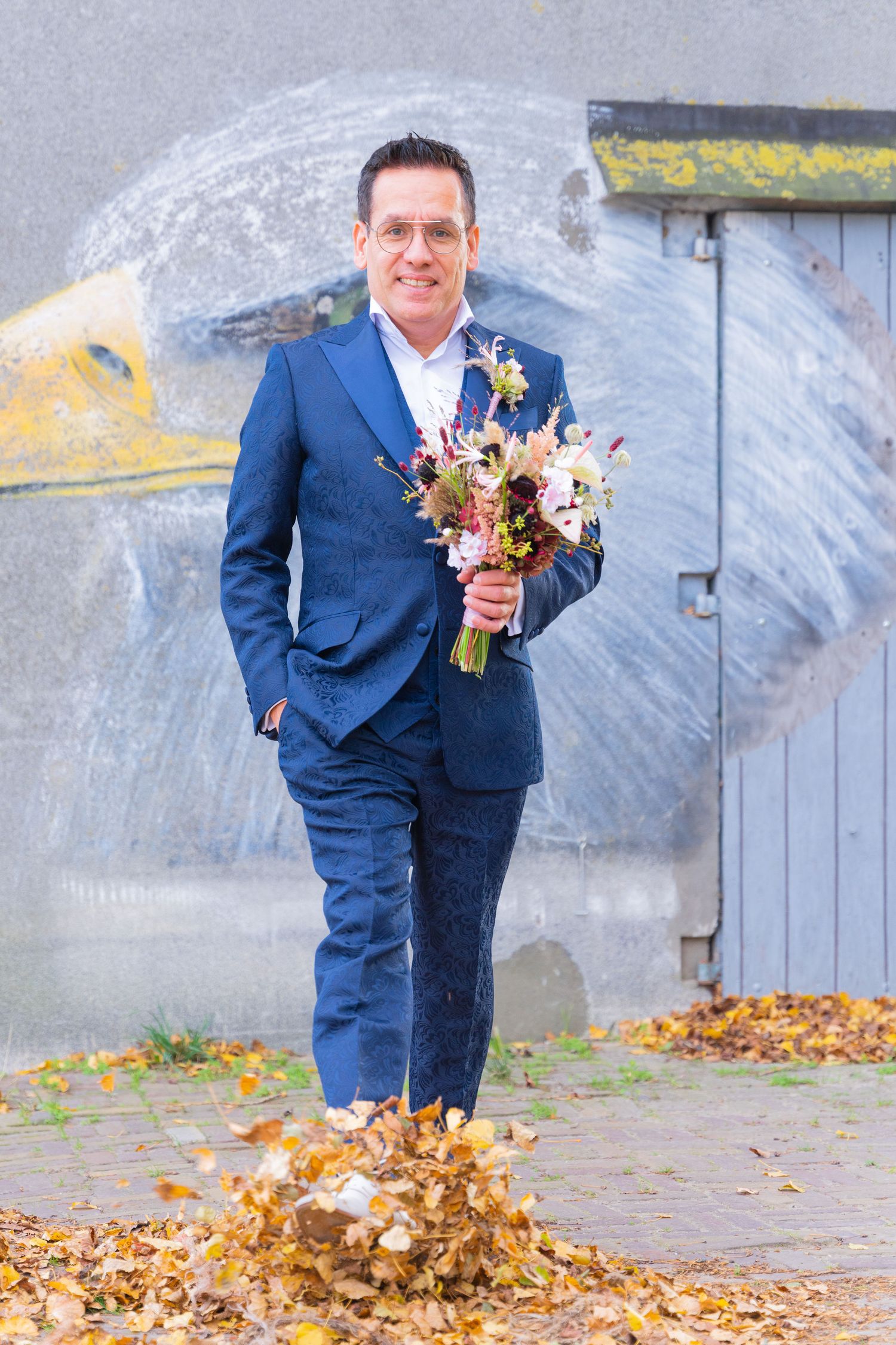 Person in blue suit playfully jumping through autumn leaves while holding a colorful bouquet.
