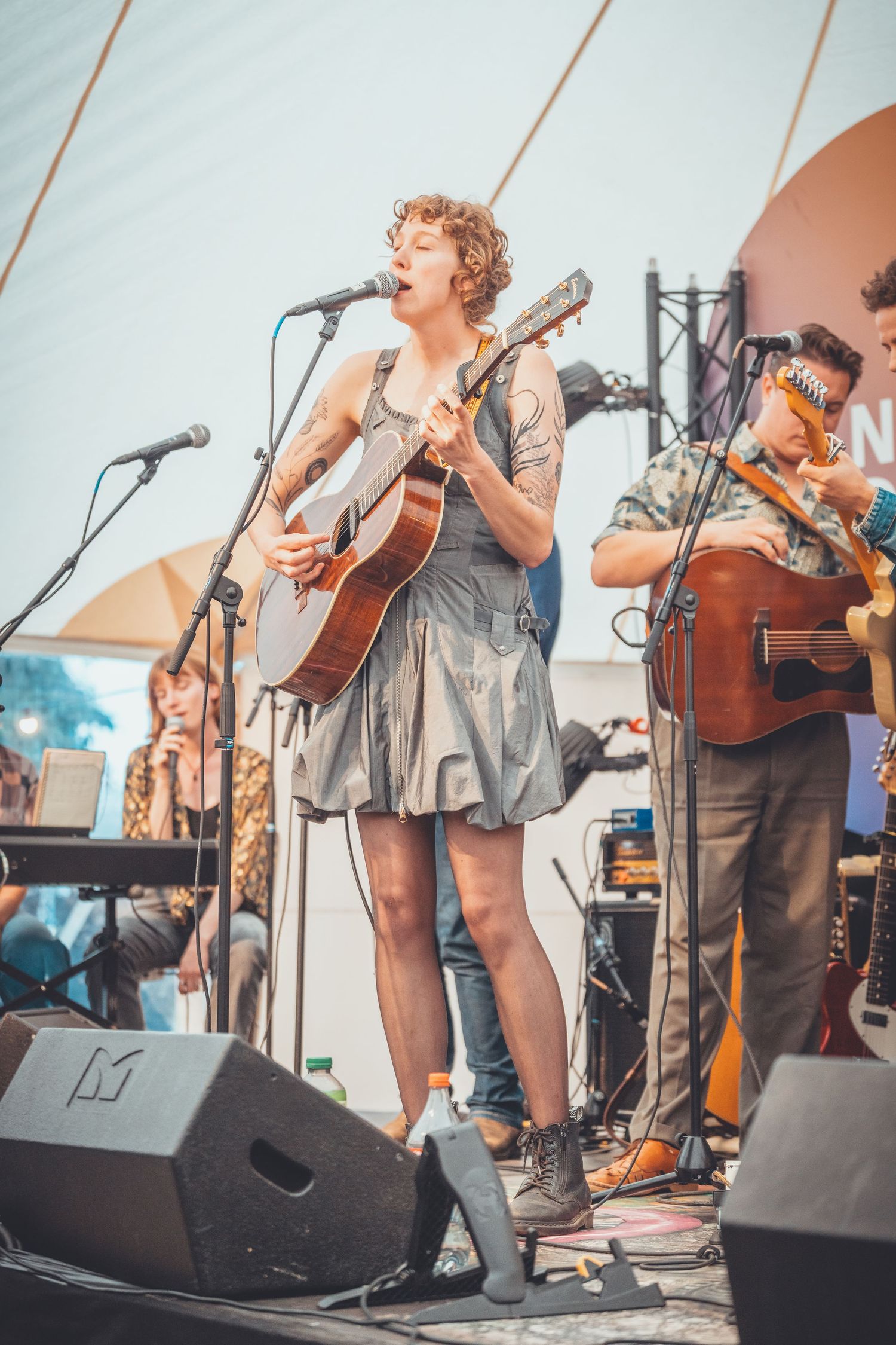Musicians perform on stage with acoustic guitars at an outdoor festival under bright lighting.