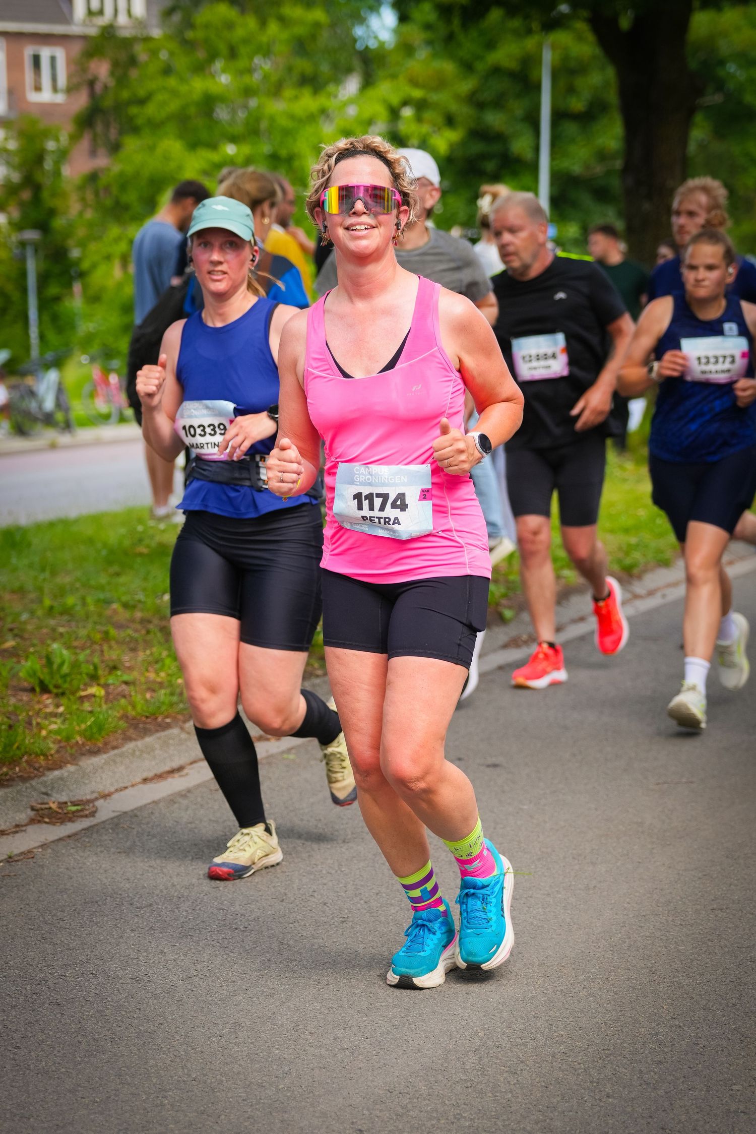 Smiling woman in pink top with bib 1174 running a road race with other participants in summer.