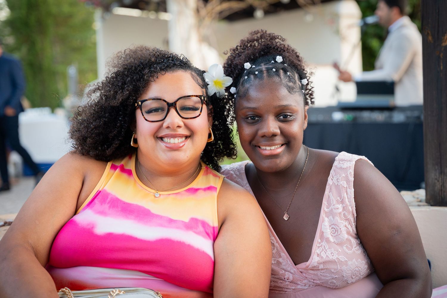 Two smiling friends sitting together at an outdoor event, one wearing a pink top and glasses, the other in pale pink lace.