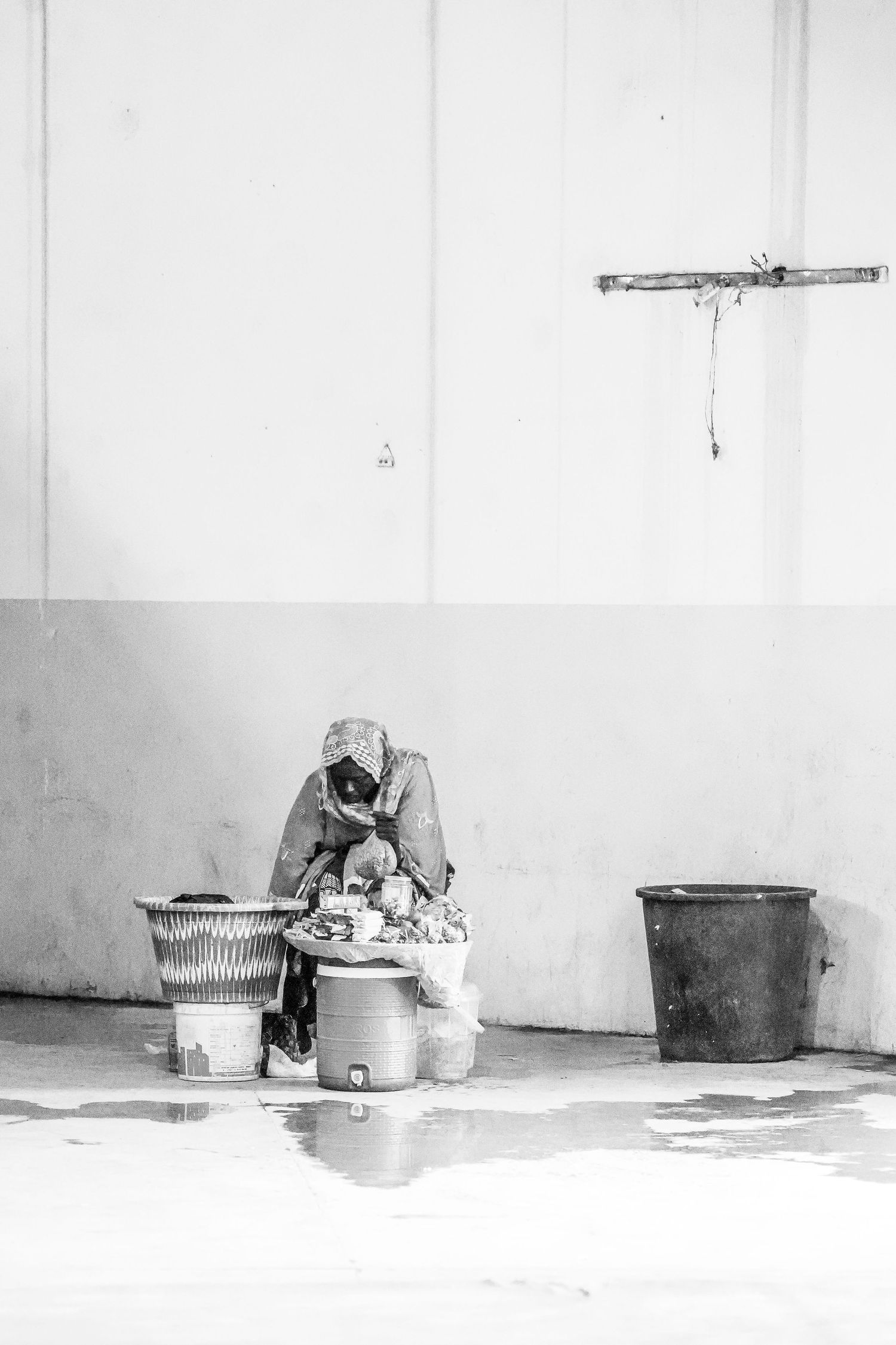 Black and white photo of a person sitting on the ground sorting goods between buckets against a white wall.