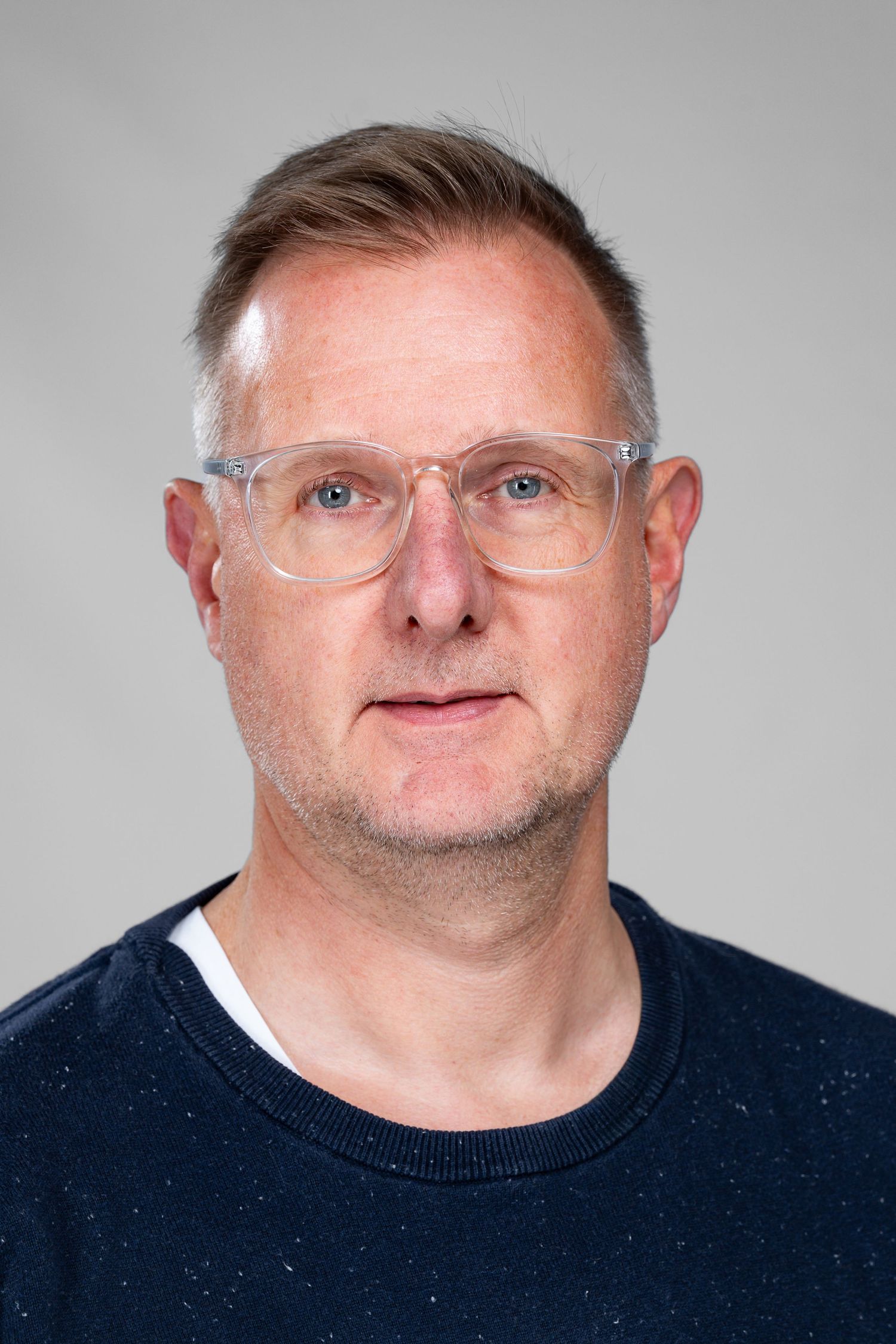 Middle-aged man with clear glasses, blue eyes, and stubble wearing a navy sweater against a gray background.