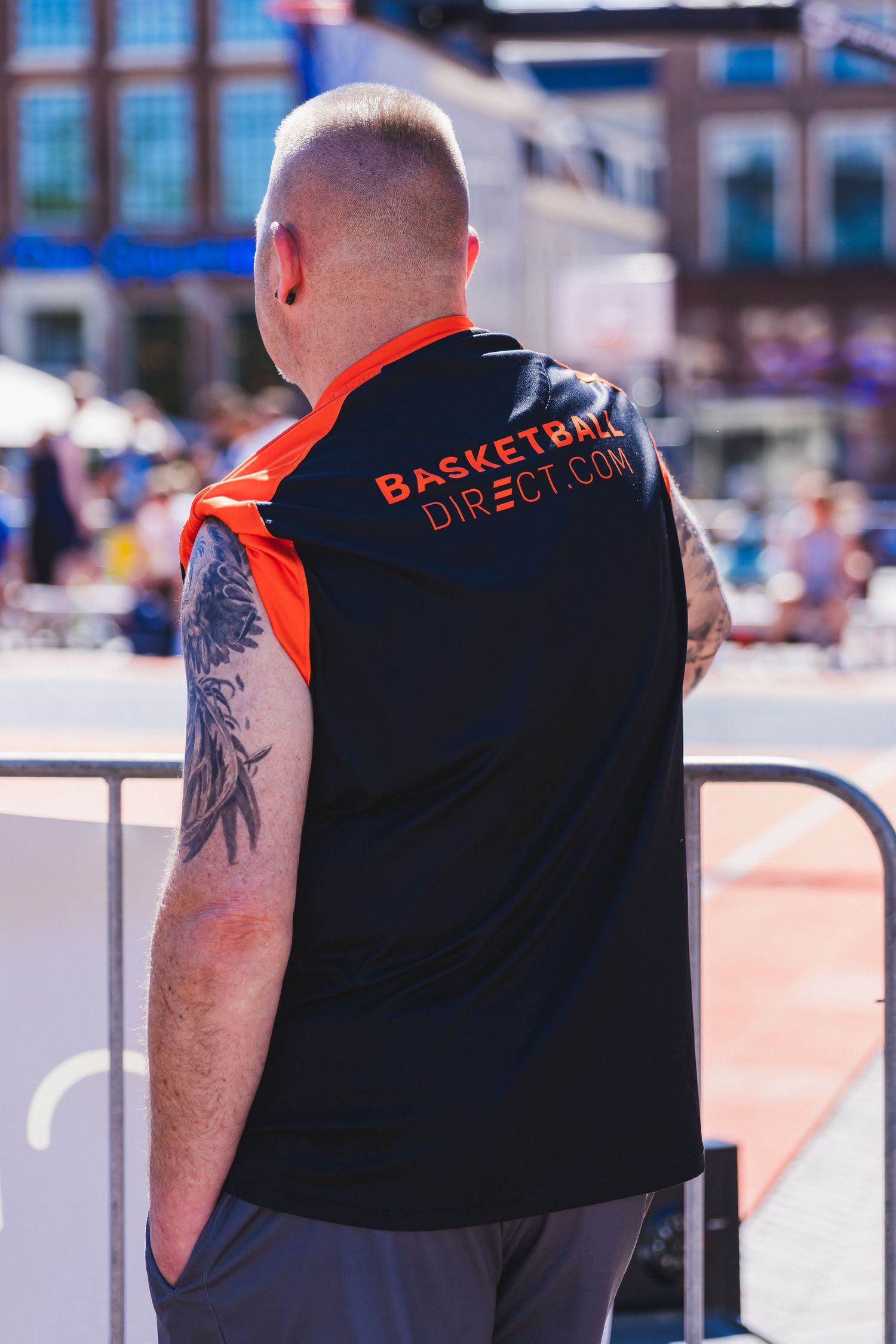 Man wearing a black and orange Basketball Direct sleeveless jersey standing outdoors at a sunny basketball event.