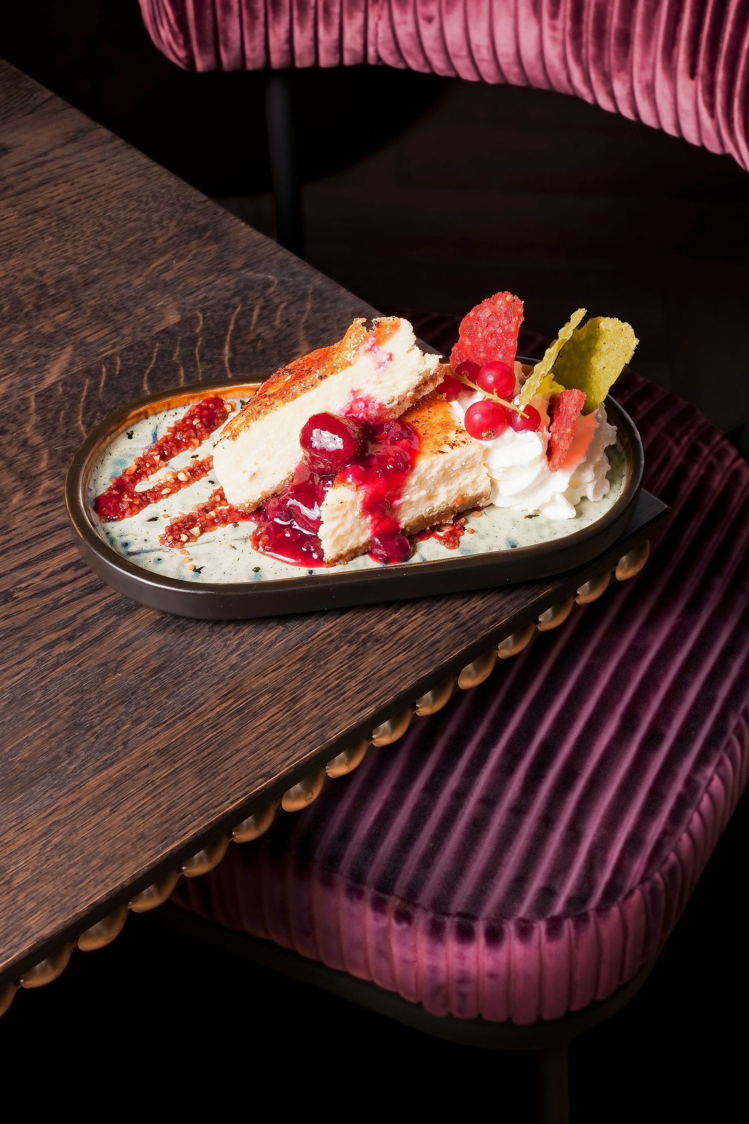 A slice of cheesecake with berry sauce and citrus garnish on an oval plate, served on a dark wooden restaurant table.