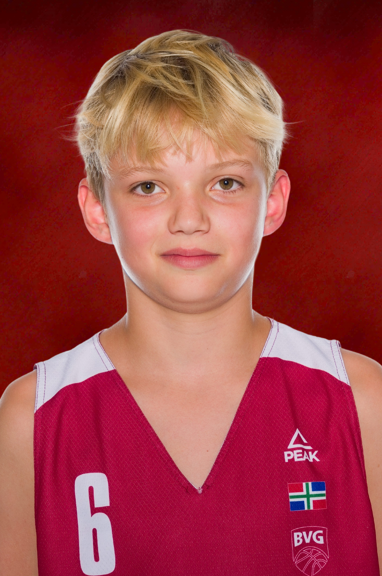Young basketball player with blond hair wearing a red number 6 Peak jersey with BVG logo, posing against a dark red backdrop.
