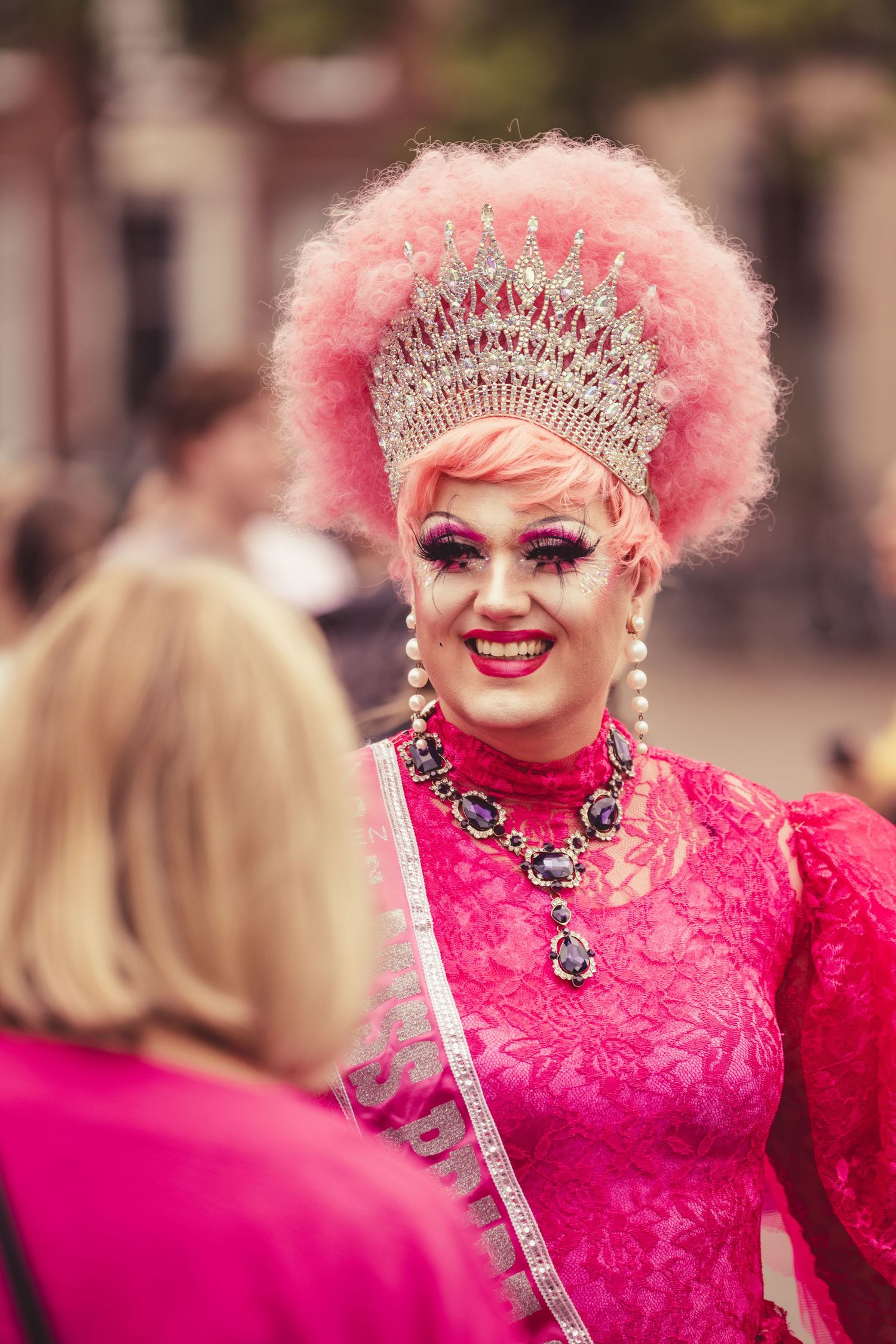 A performer in a bright pink lace dress and glittering crown poses with dramatic makeup and pink hair.