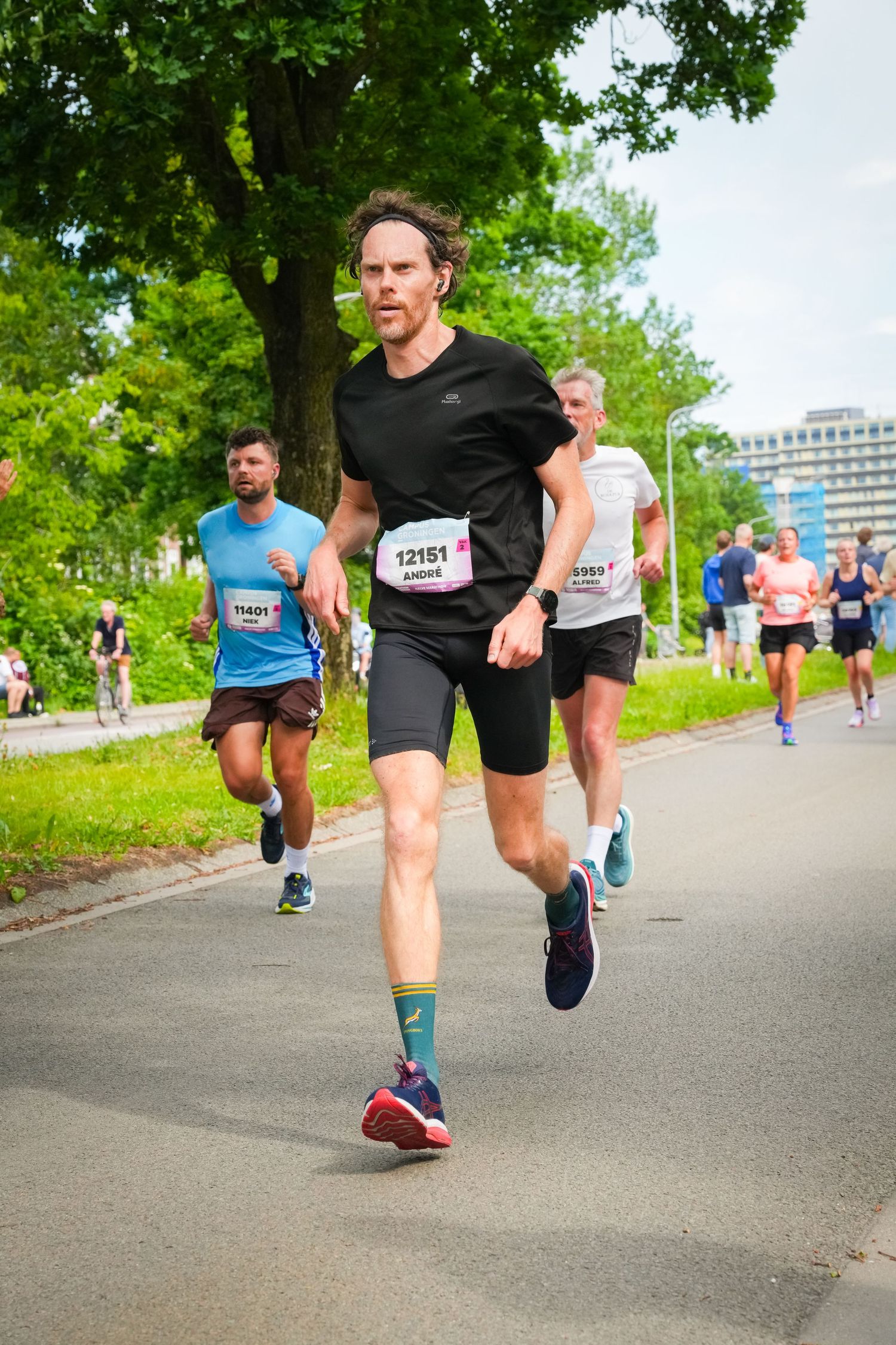 Runners competing in a road race, with bib number 12151 leading the pack through a tree-lined course.