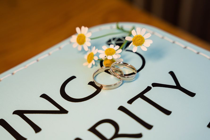 Wedding rings rest on a light blue surface with scattered white and yellow daisies creating a romantic backdrop.