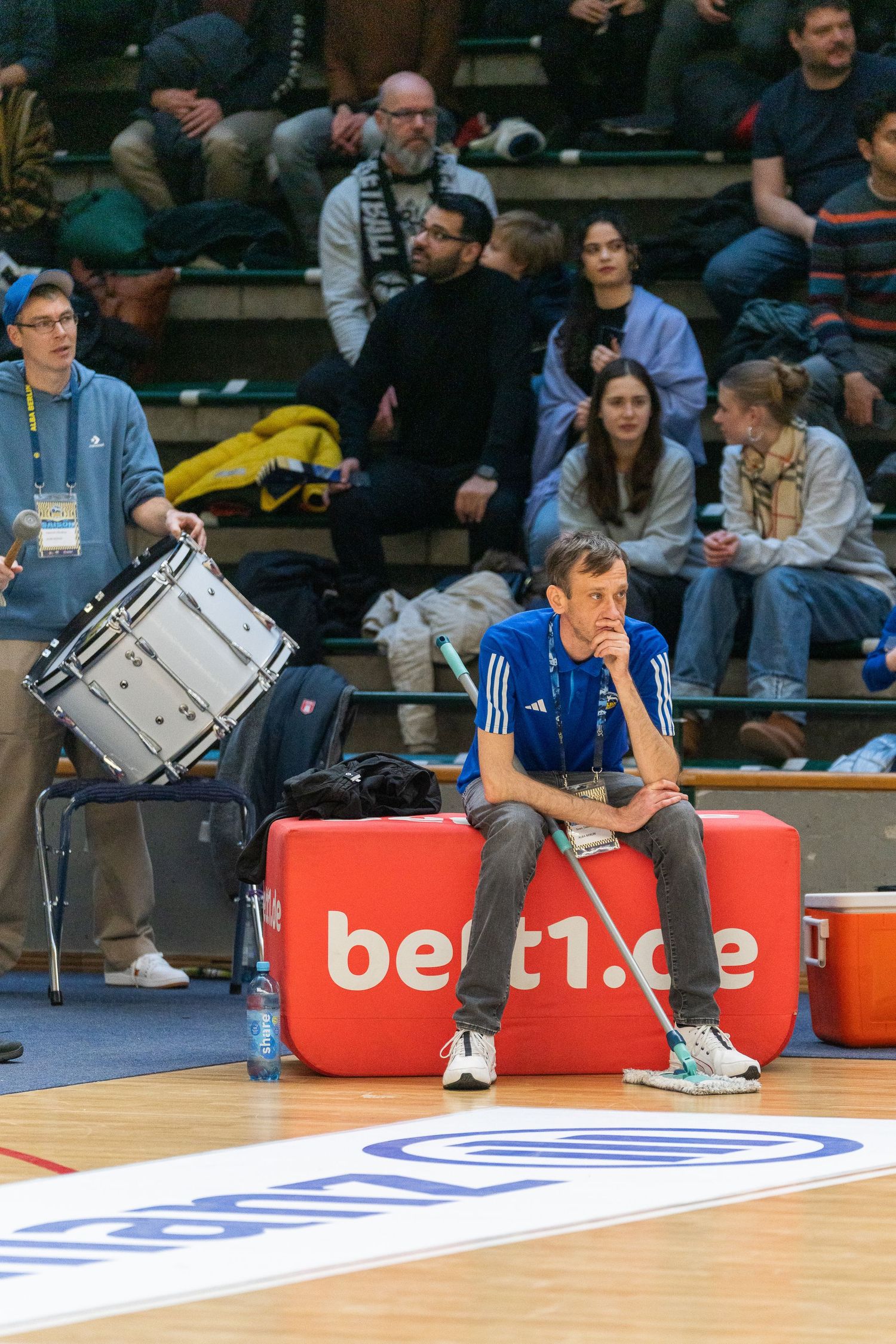 Man sitting courtside at basketball game near a snare drum, with spectators in bleachers behind him.