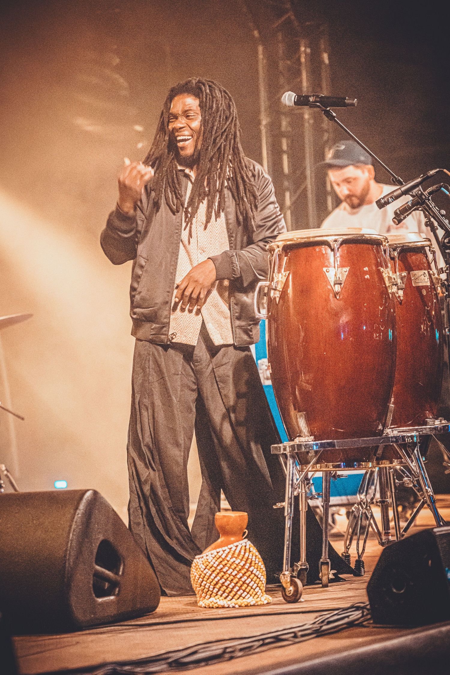 Performer with dreadlocks in black and white outfit energetically singing on stage with drummer and conga drums in background.