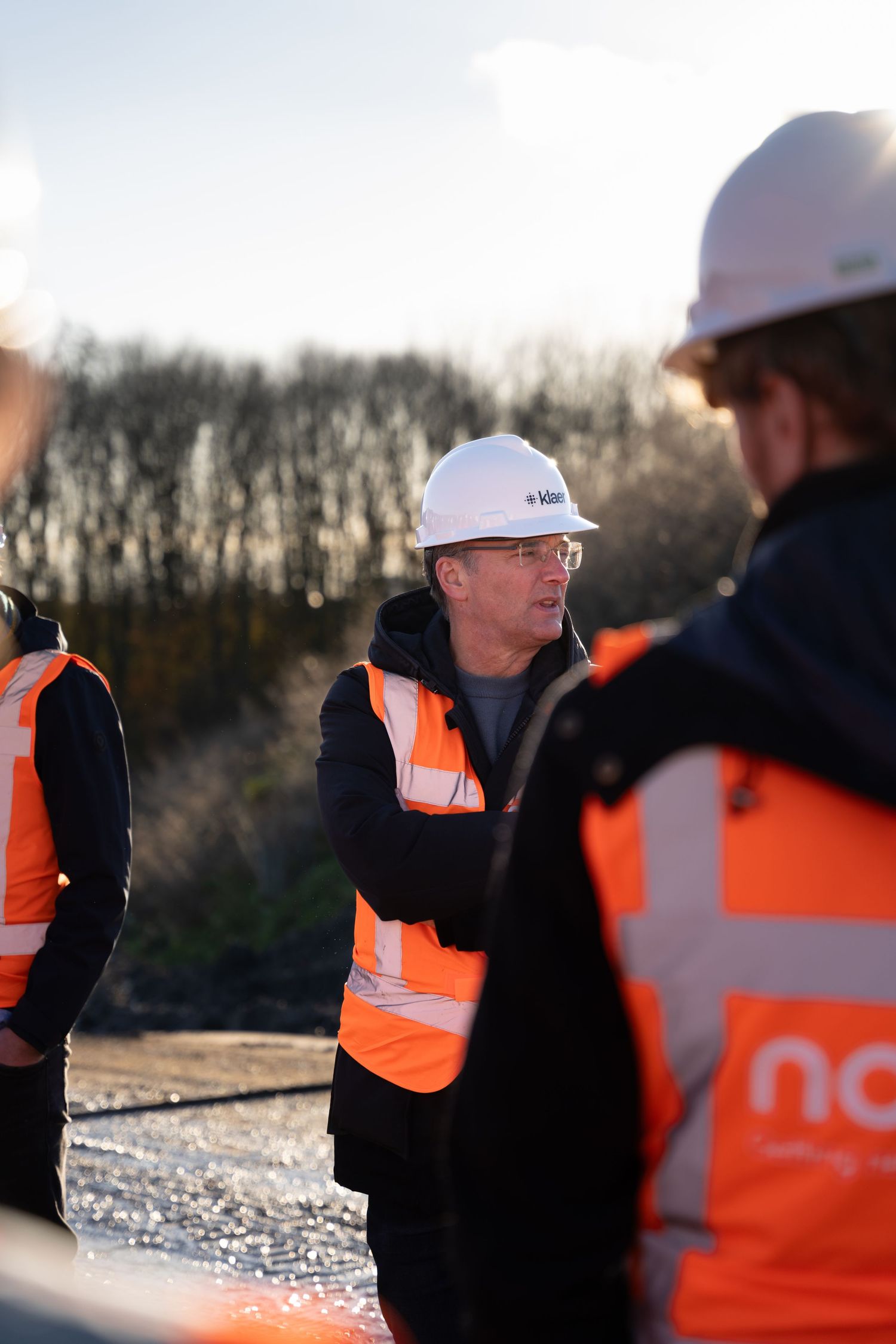Workers in orange safety vests and hard hats gather at a construction site during sunset.
