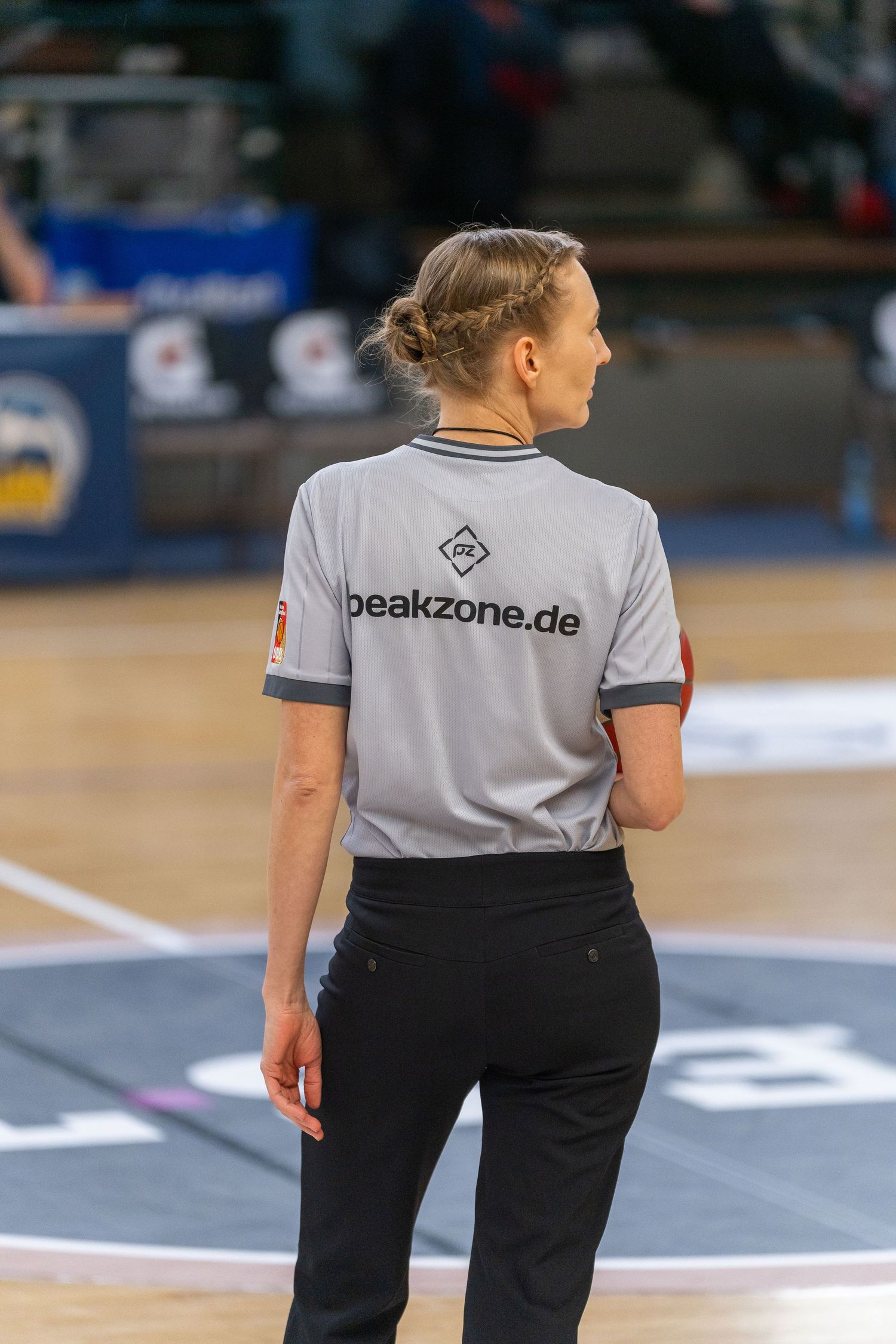 Basketball referee in grey shirt with peakzone.de branding and black pants stands on court during game.