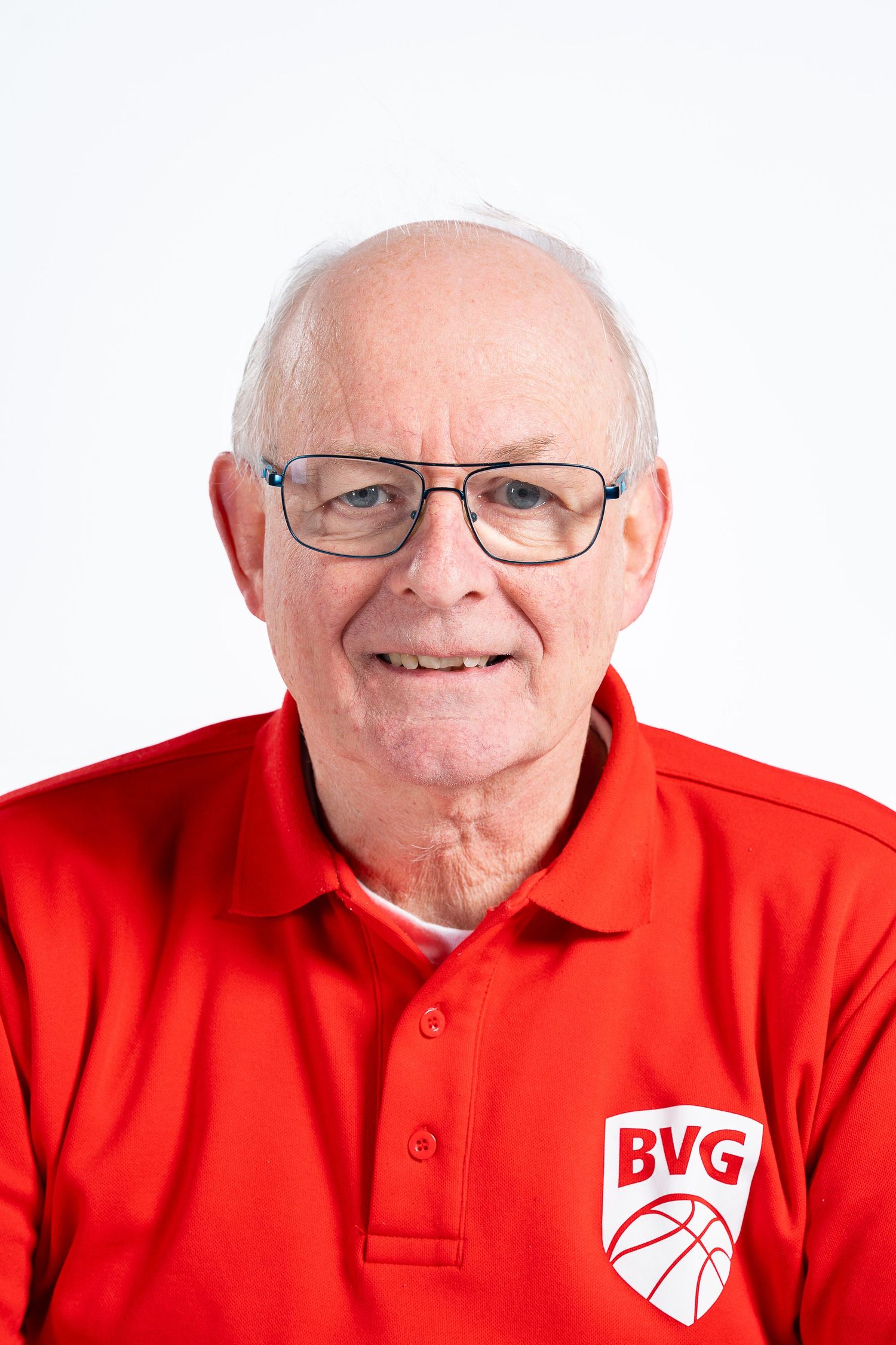 Elderly man in red BVG basketball polo shirt and glasses smiling for a portrait photo against white background.