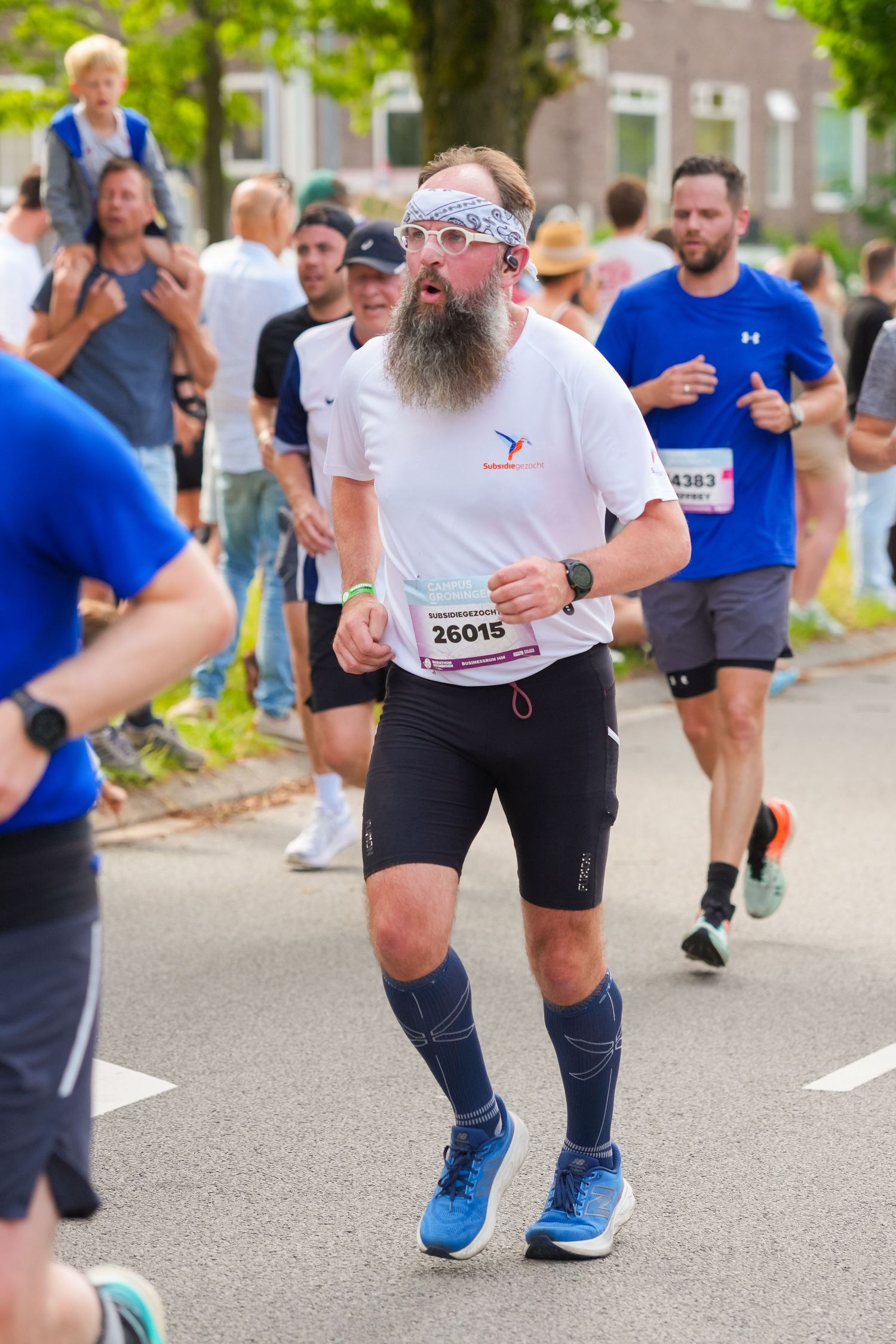 Bearded male marathon runner with bib number 26015 competing in a road race, wearing white shirt and compression socks.