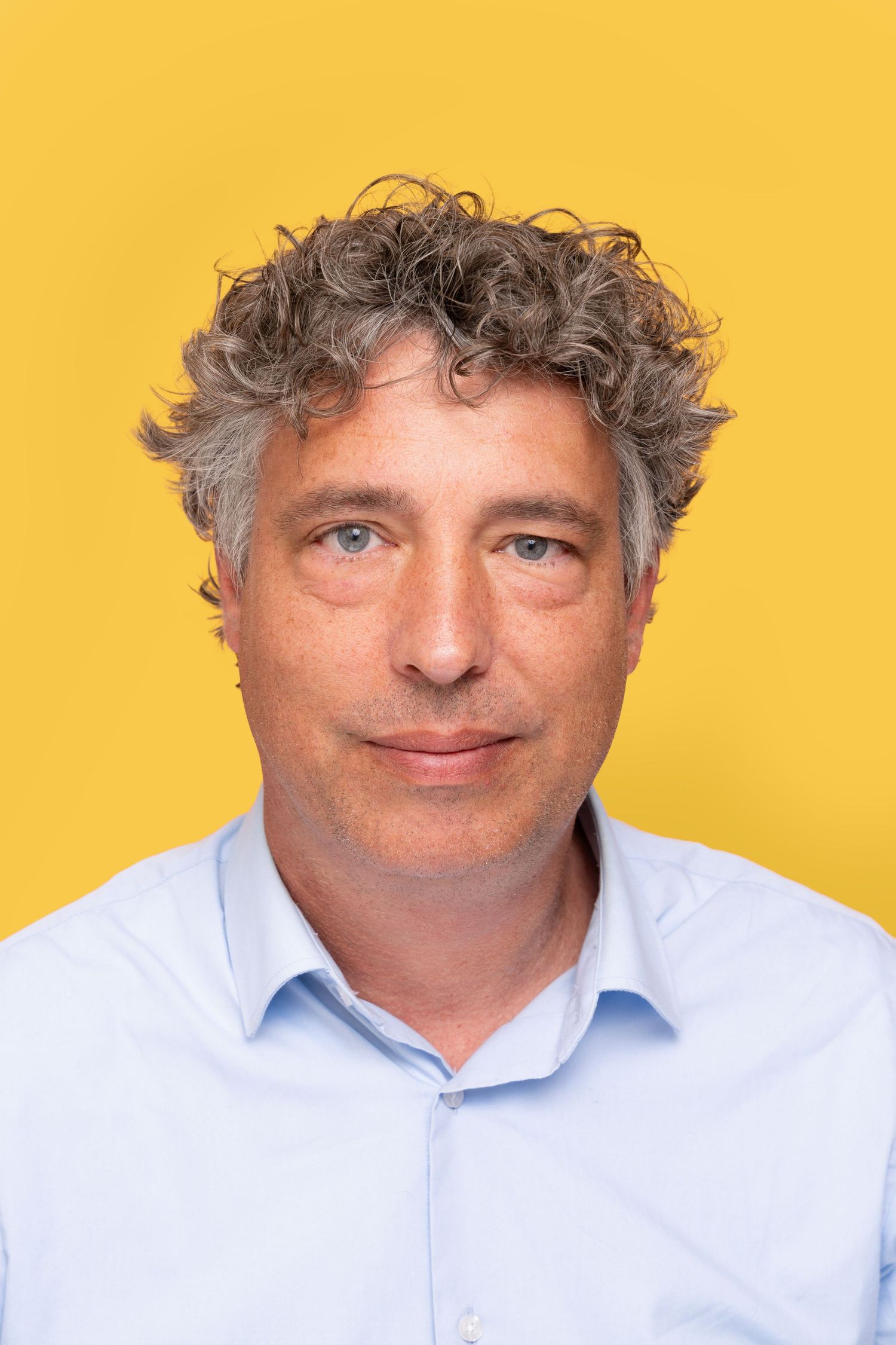 Professional headshot of a middle-aged person with curly gray hair wearing a light blue button-up shirt against a yellow background.