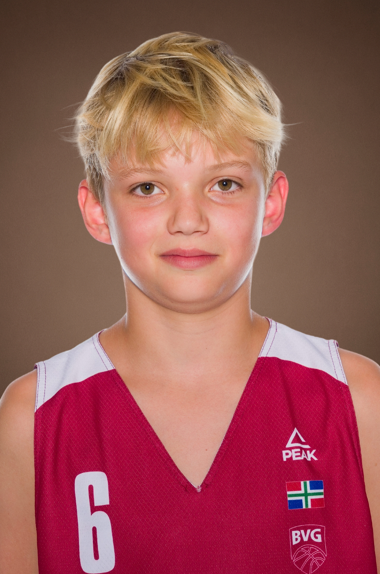 Young basketball player with blond hair in a red number 6 BVG jersey with Peak logo posing for a team portrait against a brown background.
