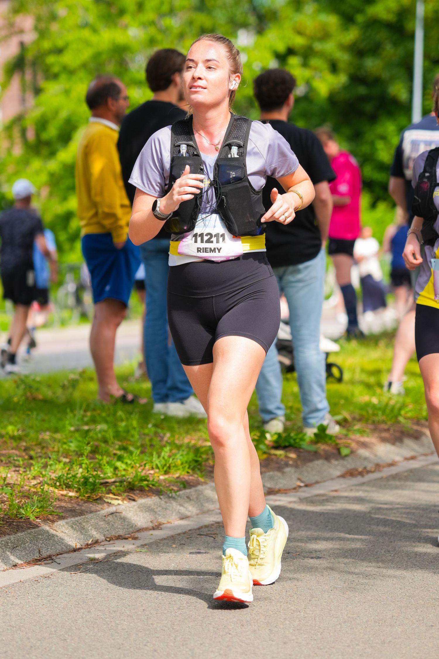 Female marathon runner wearing bib number 11211 competing in outdoor race, dressed in black shorts and hydration vest.