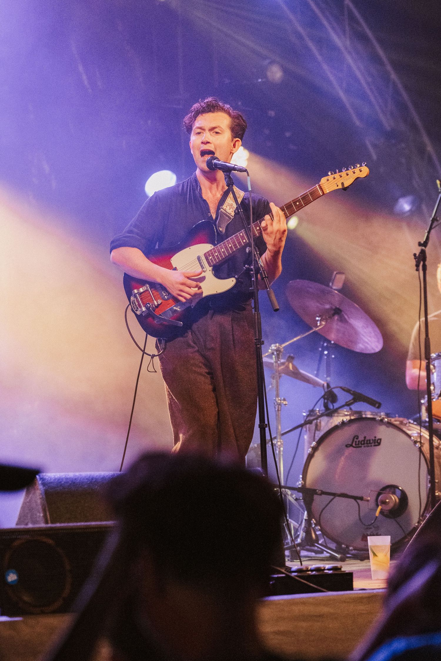 Musician performs energetically on stage with electric guitar under purple lighting while a drum kit is visible in background.