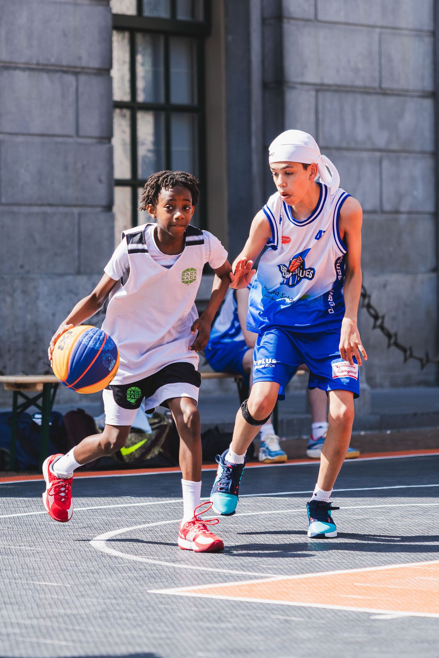Two young boys compete in street basketball, one dribbling while the other defends on an outdoor urban court.