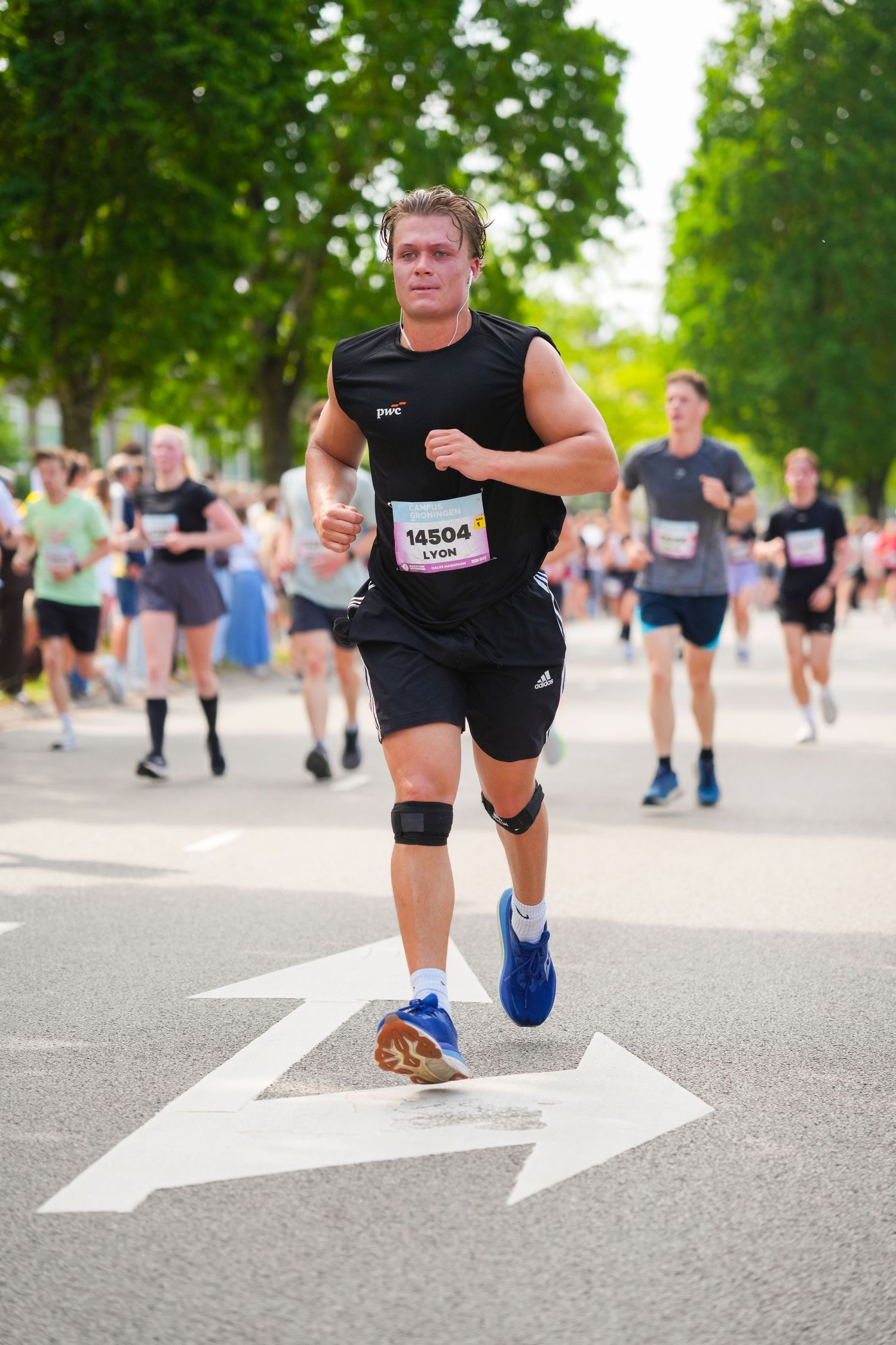 Runner #14504 from Lyon competing in a road race, wearing black athletic gear and a knee brace on a tree-lined street.